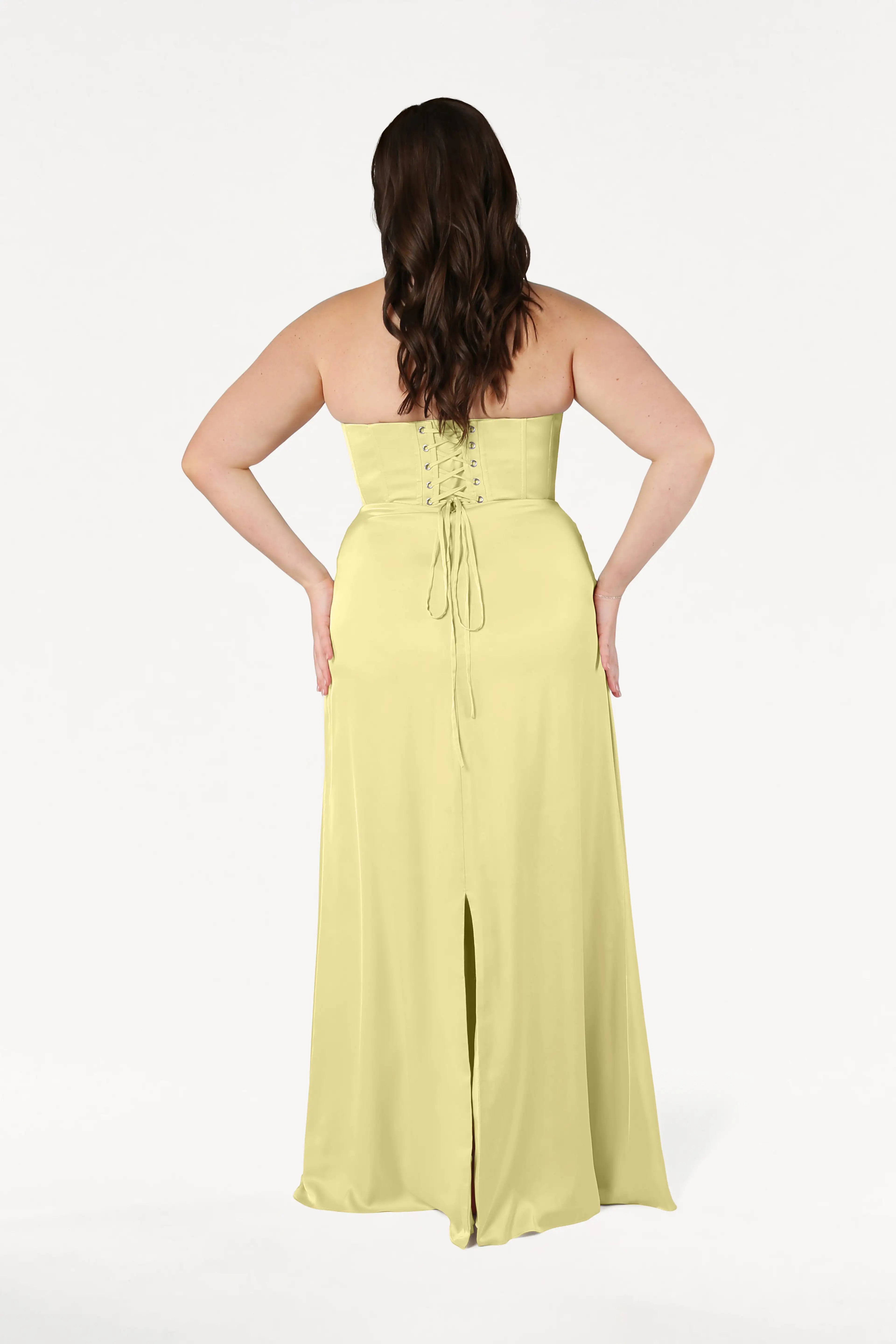 A woman with long brown hair stands with her hands on her hips, wearing the size-inclusive TH&TH Selena Bridesmaid Dress in Limoncello Satin—a strapless gown with a lace-up corset bodice and back slit—against a plain white background.