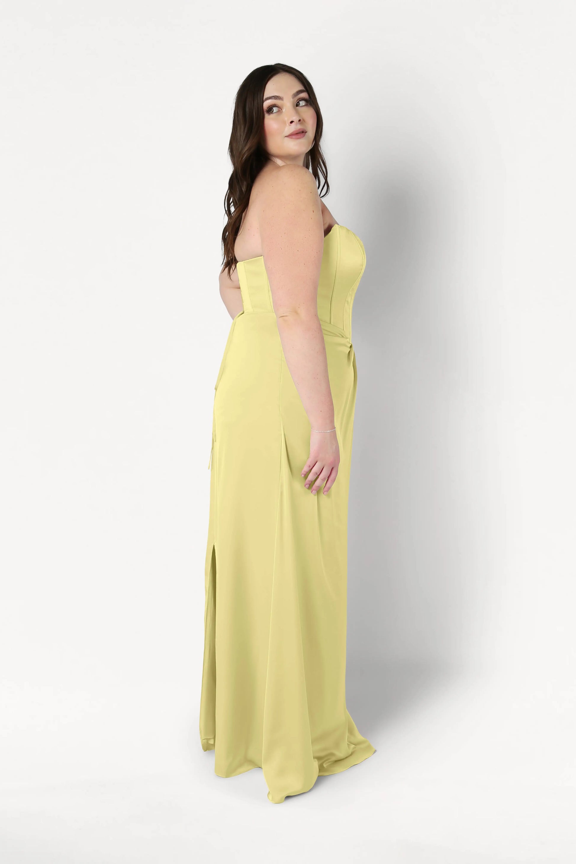 A woman with long brown hair models the TH&TH Selena Bridesmaid Dress in Limoncello Satin—a strapless, corset-bodice gown. She stands sideways with a neutral expression, looking slightly upward against a plain white background.