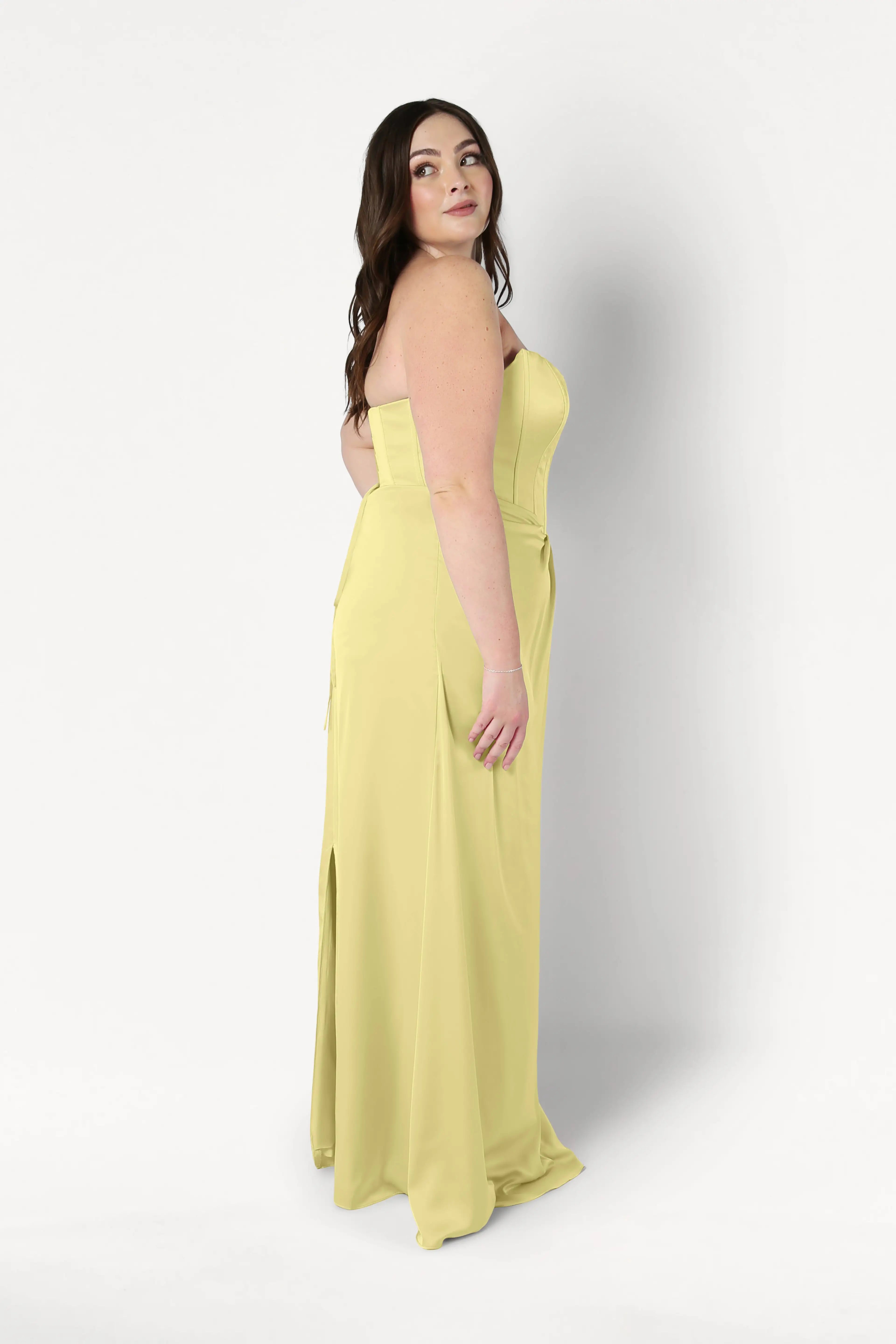 A woman with long brown hair models the TH&TH Selena Bridesmaid Dress in Limoncello Satin—a strapless, corset-bodice gown. She stands sideways with a neutral expression, looking slightly upward against a plain white background.