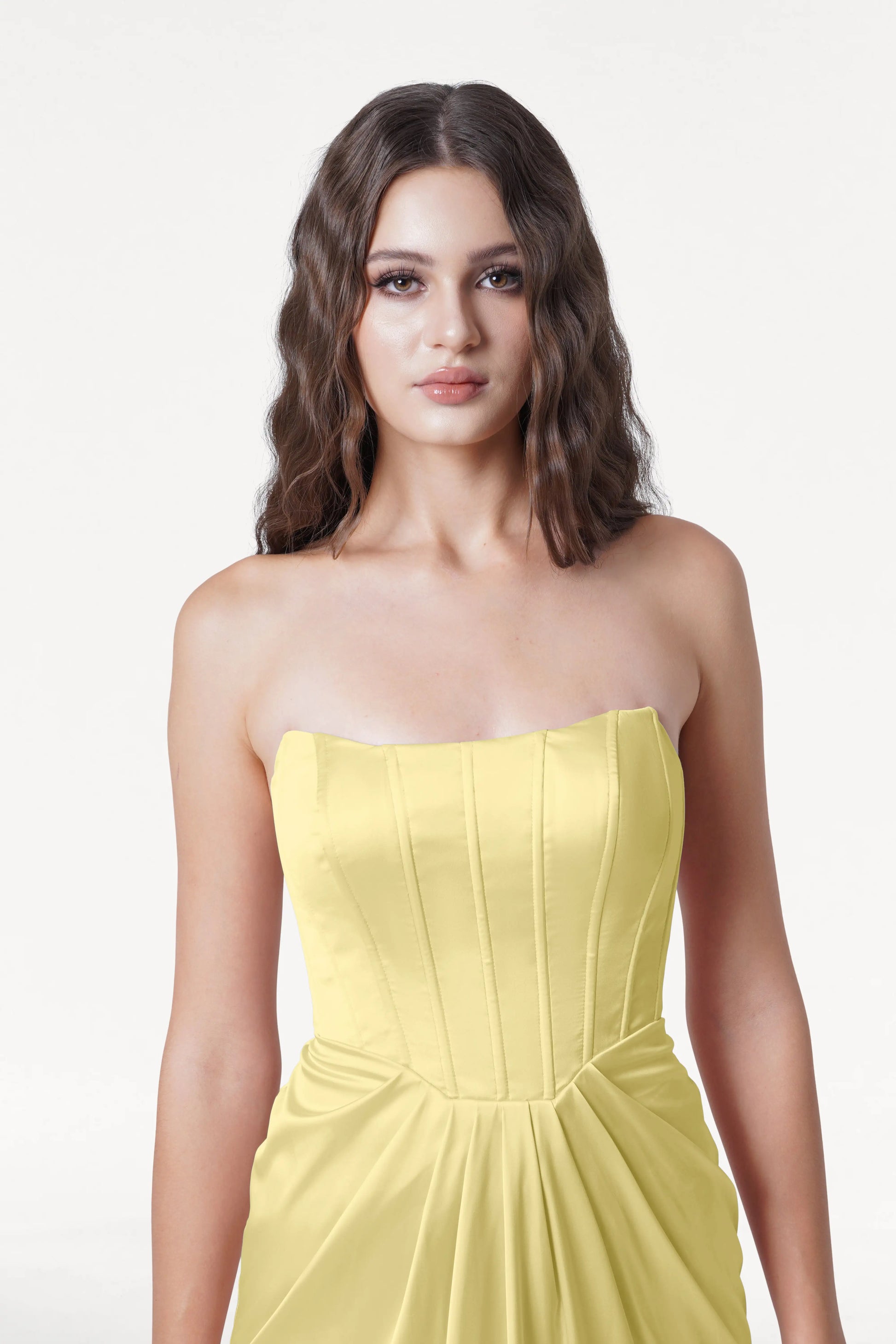 A woman with wavy brown hair wears the Selena Bridesmaid Dress in Limoncello Satin by TH&TH, featuring a strapless fitted corset bodice and pleated skirt, against a plain white background.