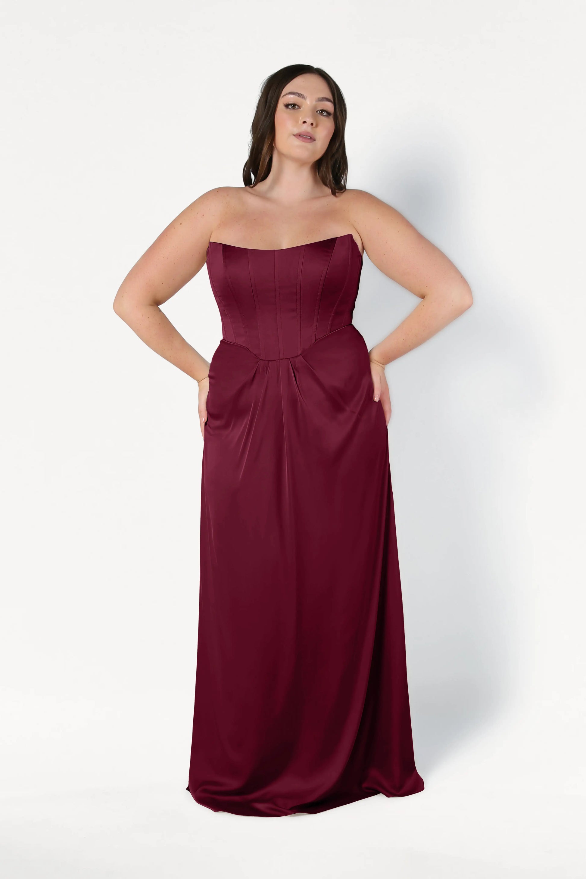 A woman wears the TH&TH Selena Bridesmaid Dress in Merlot Satin—a size-inclusive, strapless, floor-length gown with a fitted corset bodice and flowing skirt—standing against a white background with her hands in the dress pockets.