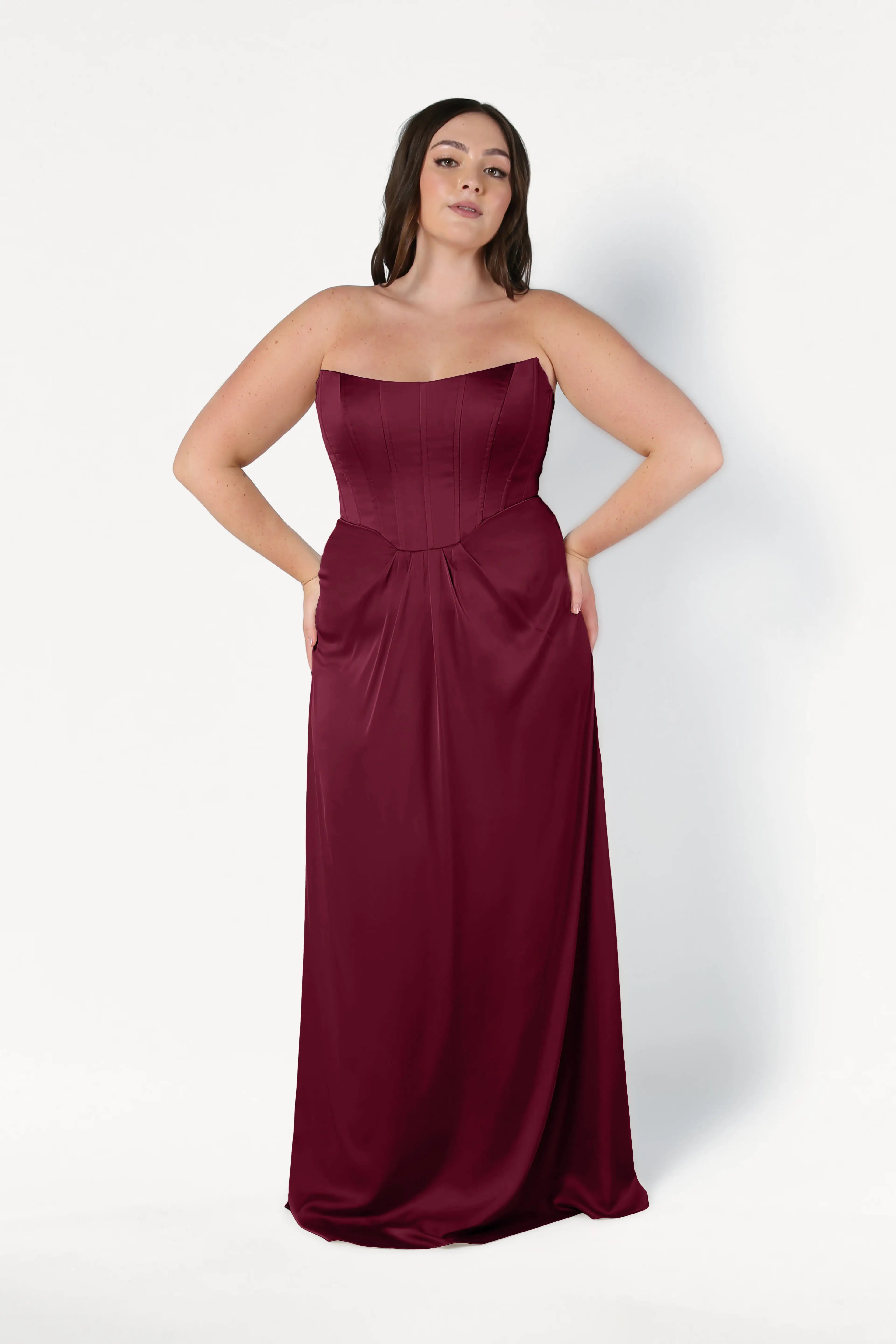 A woman wears the TH&TH Selena Bridesmaid Dress in Merlot Satin—a size-inclusive, strapless, floor-length gown with a fitted corset bodice and flowing skirt—standing against a white background with her hands in the dress pockets.