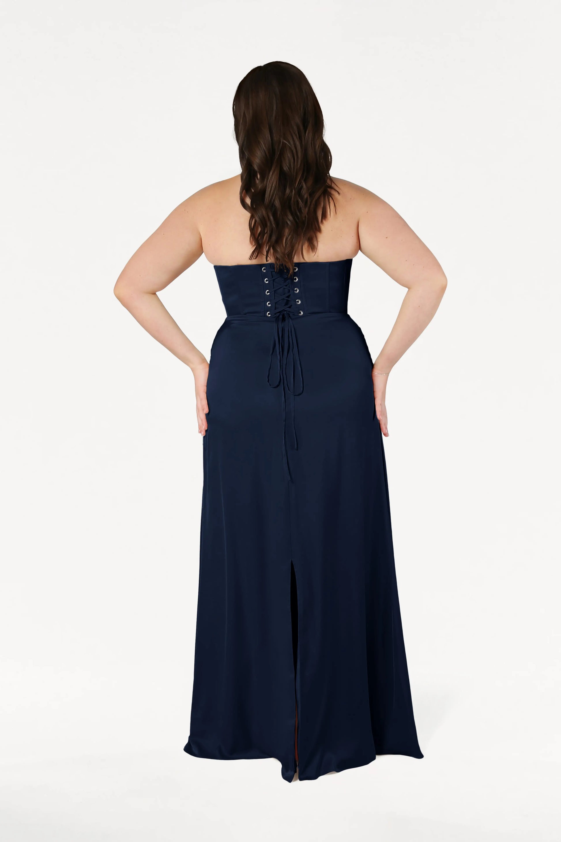 A woman is shown from the back wearing the TH&TH Selena Bridesmaid Dress in Navy Satin, featuring a strapless corset bodice with lace-up detail and back slit. She has long brown hair and poses against a white background, hands on hips.