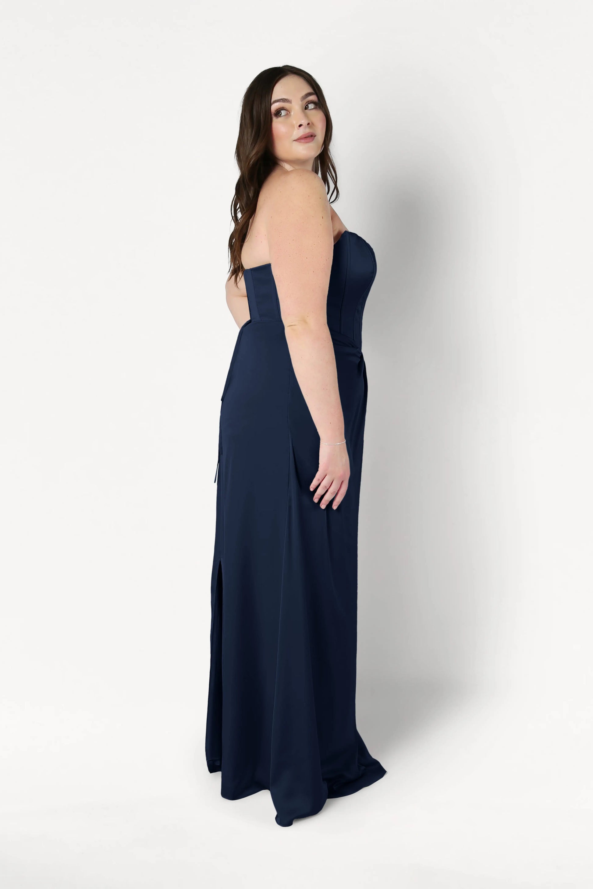 A woman with long brown hair models the TH&TH Selena Bridesmaid Dress in Navy Satin, a size-inclusive, strapless, floor-length gown. She stands sideways on a white background, gazing calmly over her shoulder.