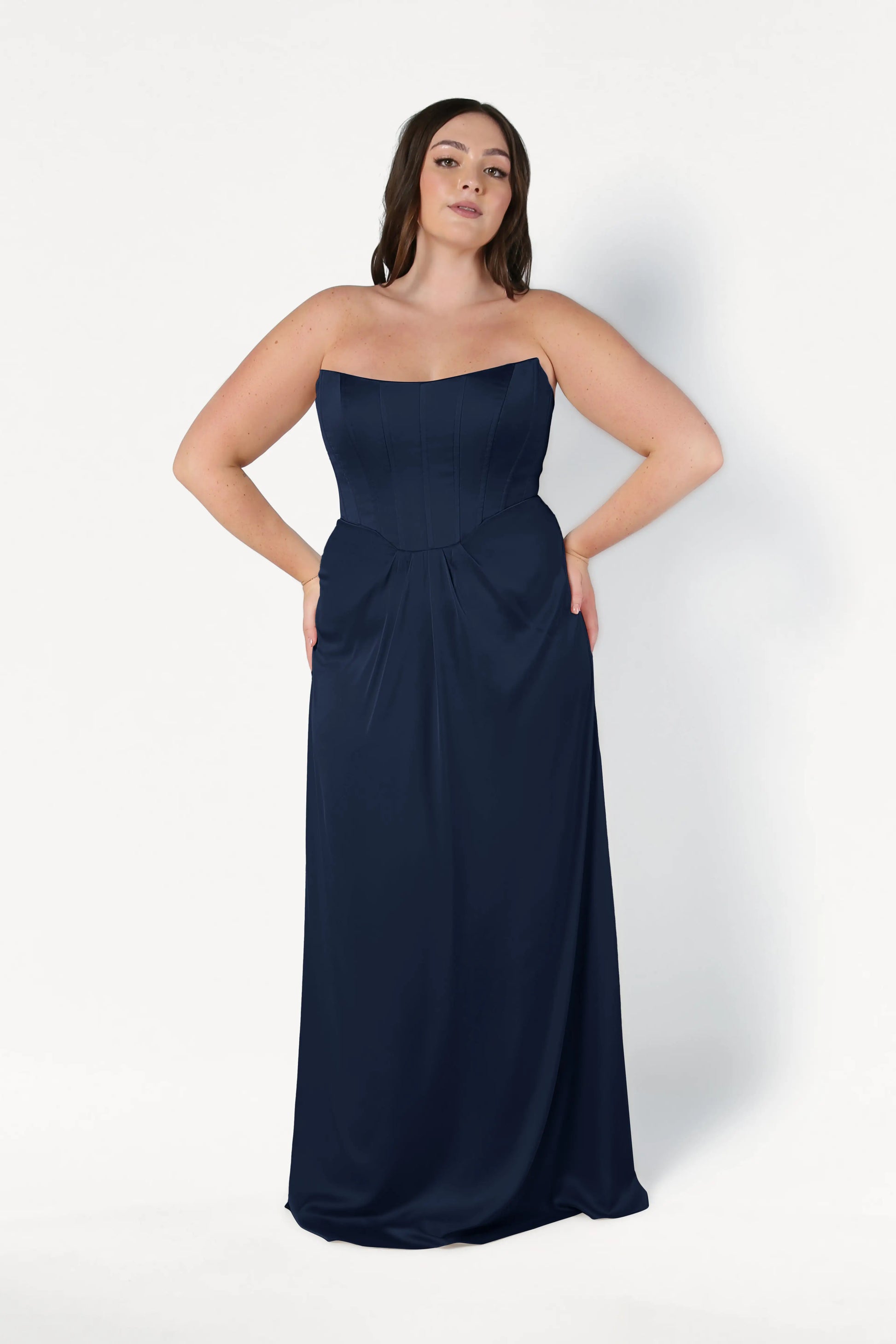 A woman wears the size-inclusive TH&TH Selena Bridesmaid Dress in Navy Satin, featuring a strapless, floor-length corset bodice, standing against a plain white background with her hands on her hips.