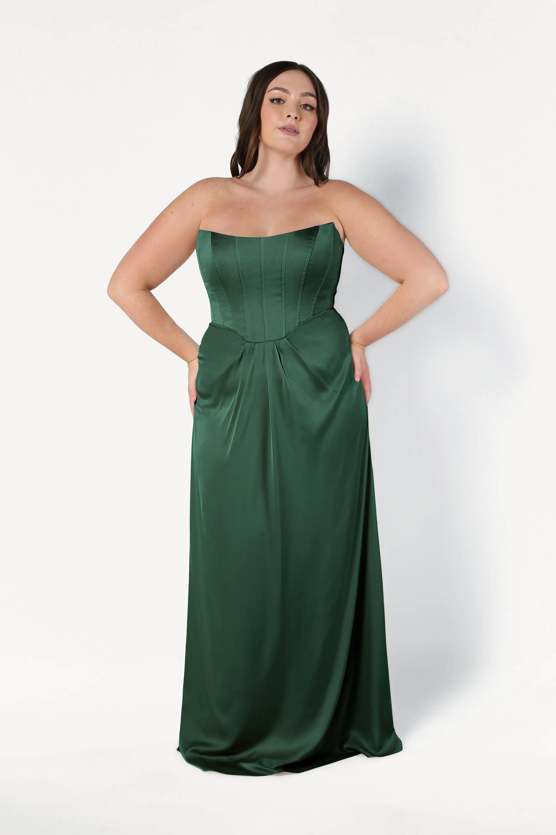 A woman confidently poses against a white background, hands on hips, wearing the TH&TH Selena Bridesmaid Dress in Olive Grove Satin—a size-inclusive, strapless, floor-length gown with a corset bodice.