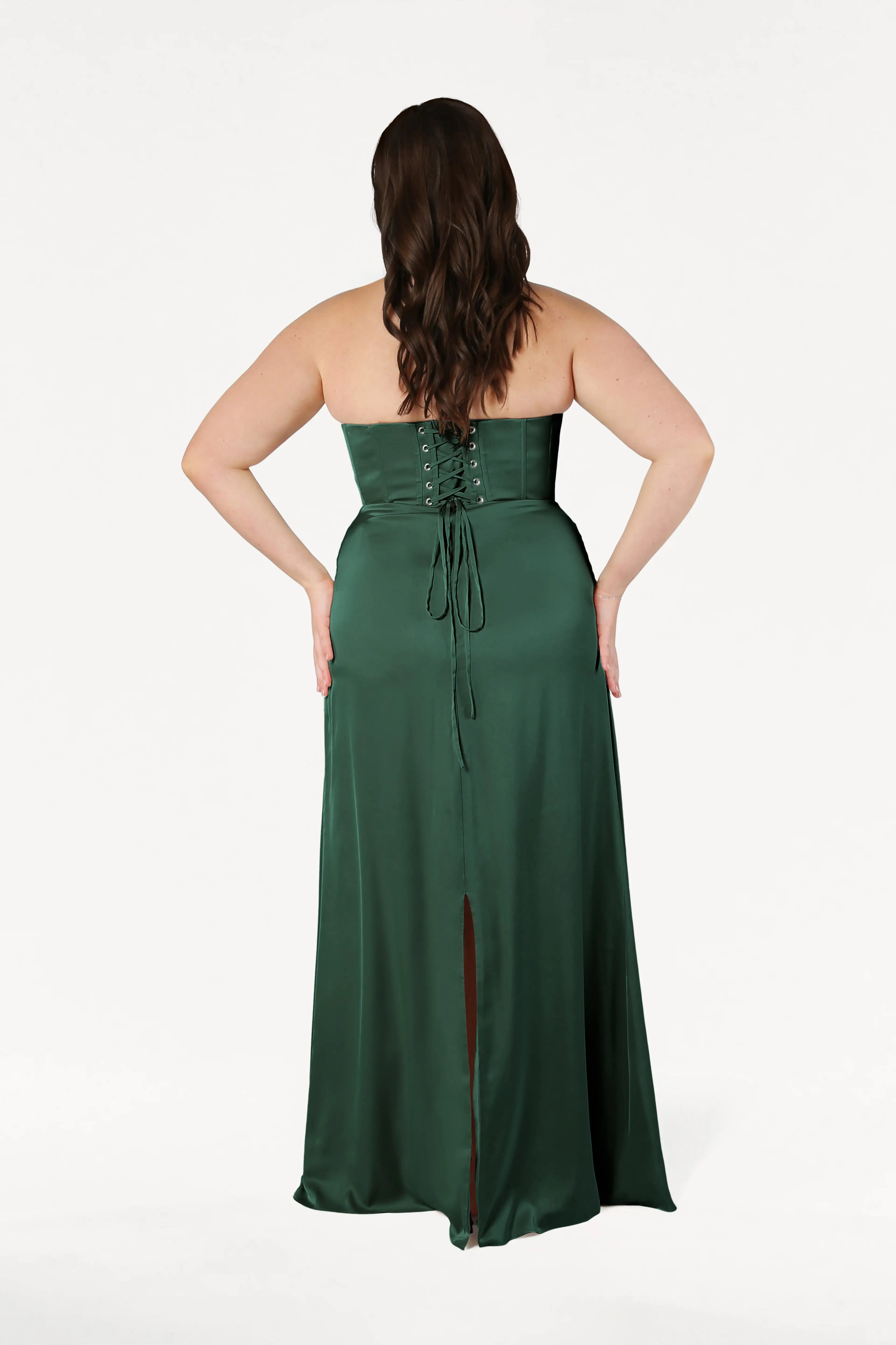 A woman with long brown hair, seen from the back, models the size-inclusive TH&TH Selena Bridesmaid Dress in Olive Grove Satin, featuring a lace-up corset bodice and skirt slit, against a plain white background.