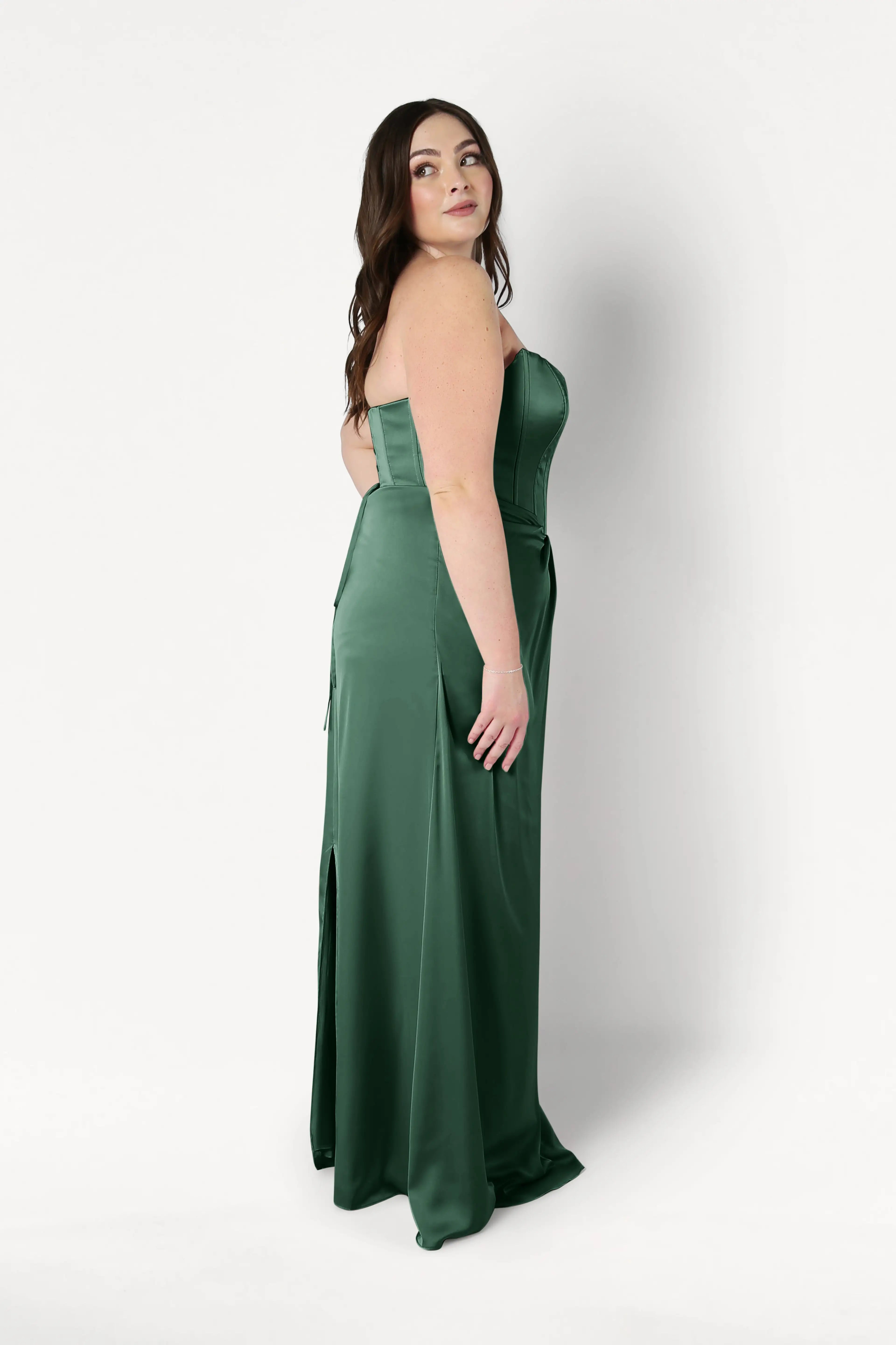 A woman with long brown hair stands sideways, wearing the size-inclusive Selena Bridesmaid Dress in Olive Grove Satin by TH&TH, featuring a floor-length silhouette and a slit. She looks over her shoulder against a plain white background.