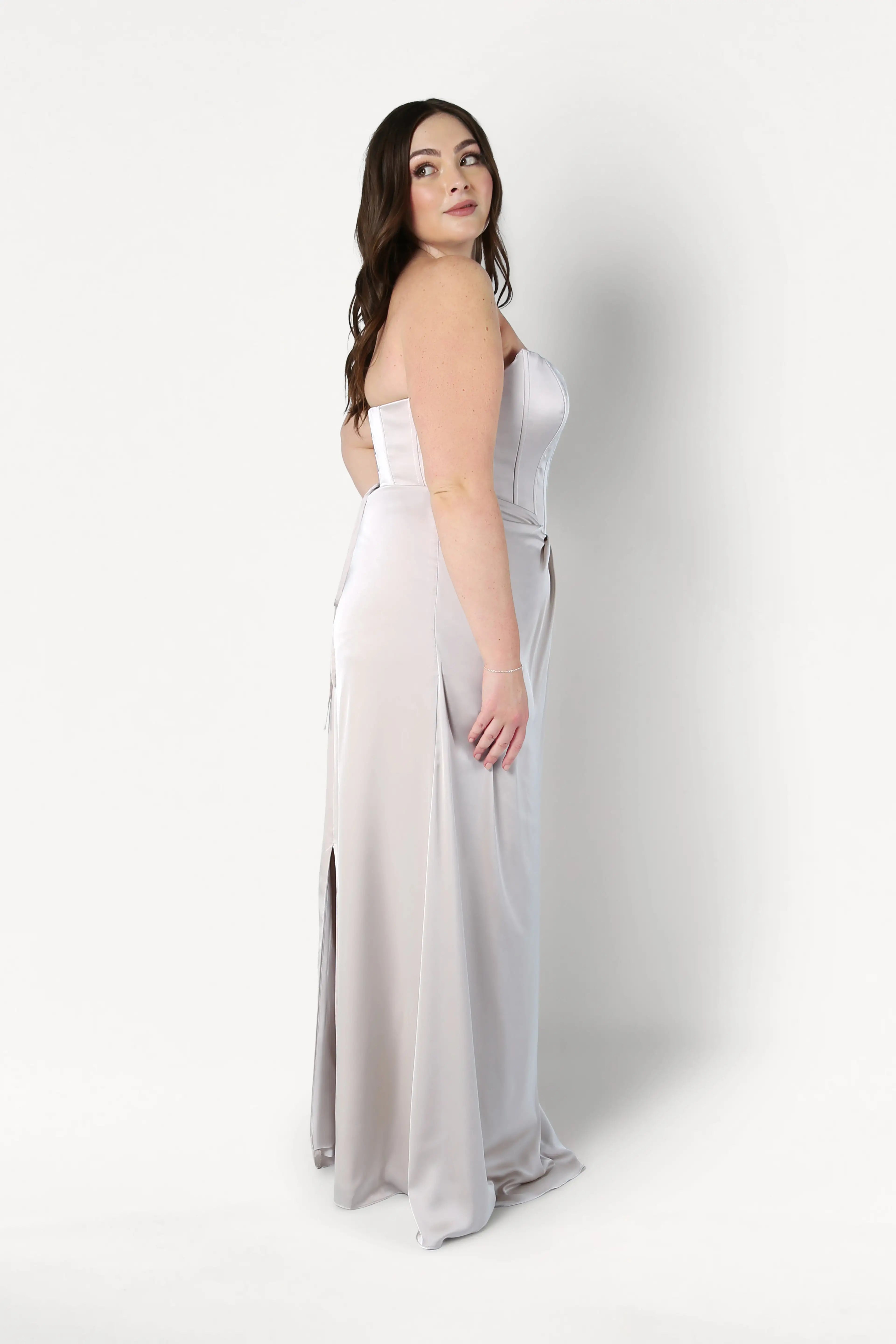 A woman with long brown hair wears the TH&TH Selena Bridesmaid Dress in Oyster Satin, featuring a corset bodice and side slit, standing in profile against a plain white background.