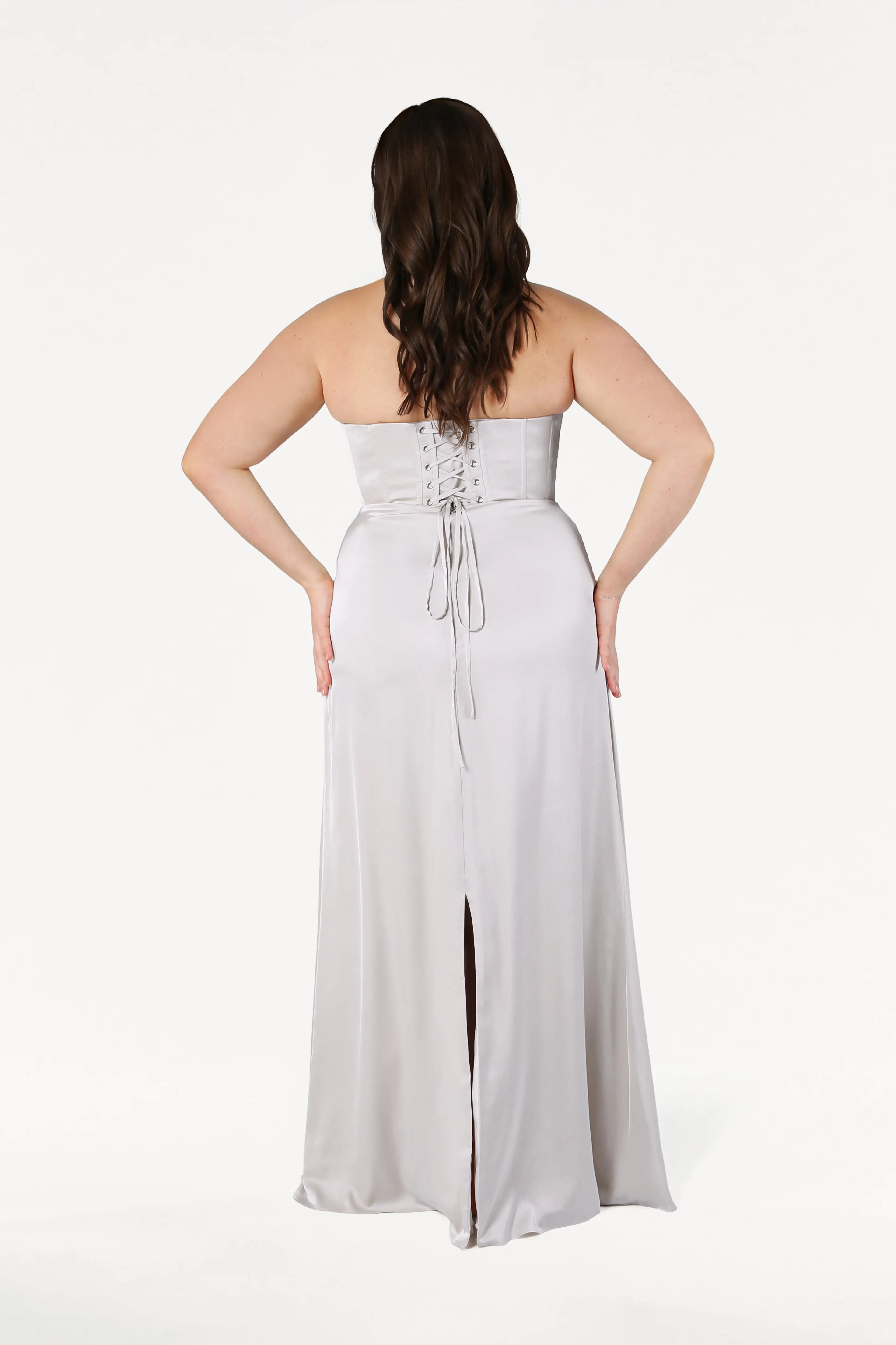 A woman with wavy brown hair models the TH&TH Selena Bridesmaid Dress in Oyster Satin, featuring a corset bodice, lace-up back, and skirt slit; the size-inclusive gown is shown from behind against a white background.