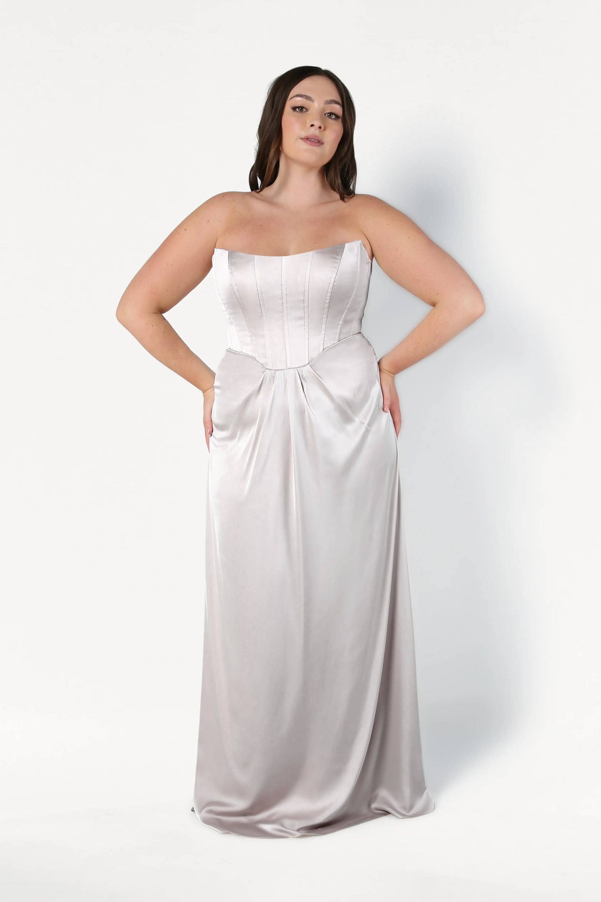A woman stands against a white background wearing the Selena Bridesmaid Dress in Oyster Satin by TH&TH, a size-inclusive, strapless, floor-length gown with a corset bodice and subtle waist gathering. She poses with hands on hips and has long, dark hair.