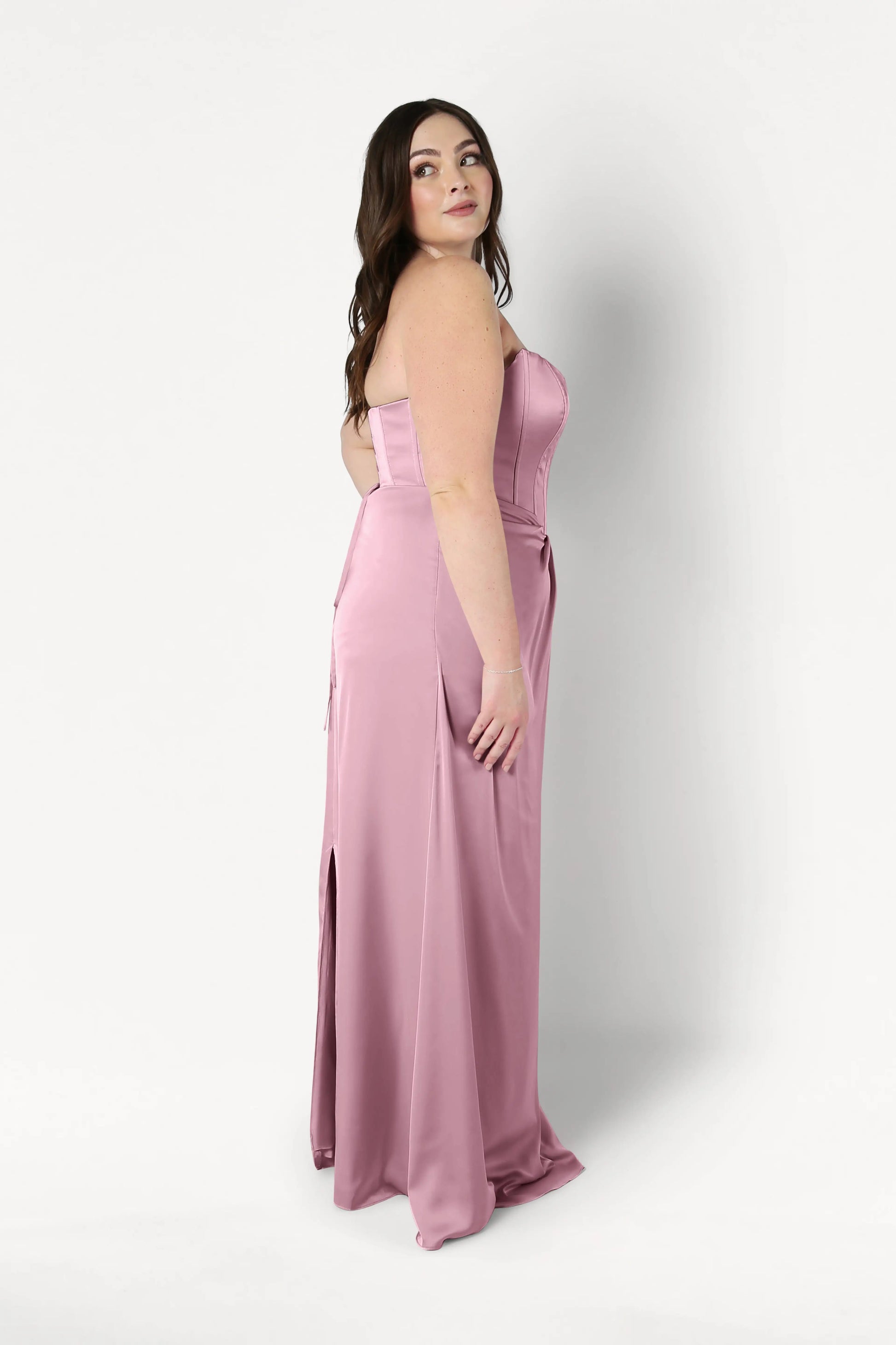 A woman stands sideways, looking over her shoulder in the Selena Bridesmaid Dress by TH&TH—a strapless, floor-length rose pink satin gown with a fitted corset bodice and slight slit—set against a white background. This style is beautifully size-inclusive.