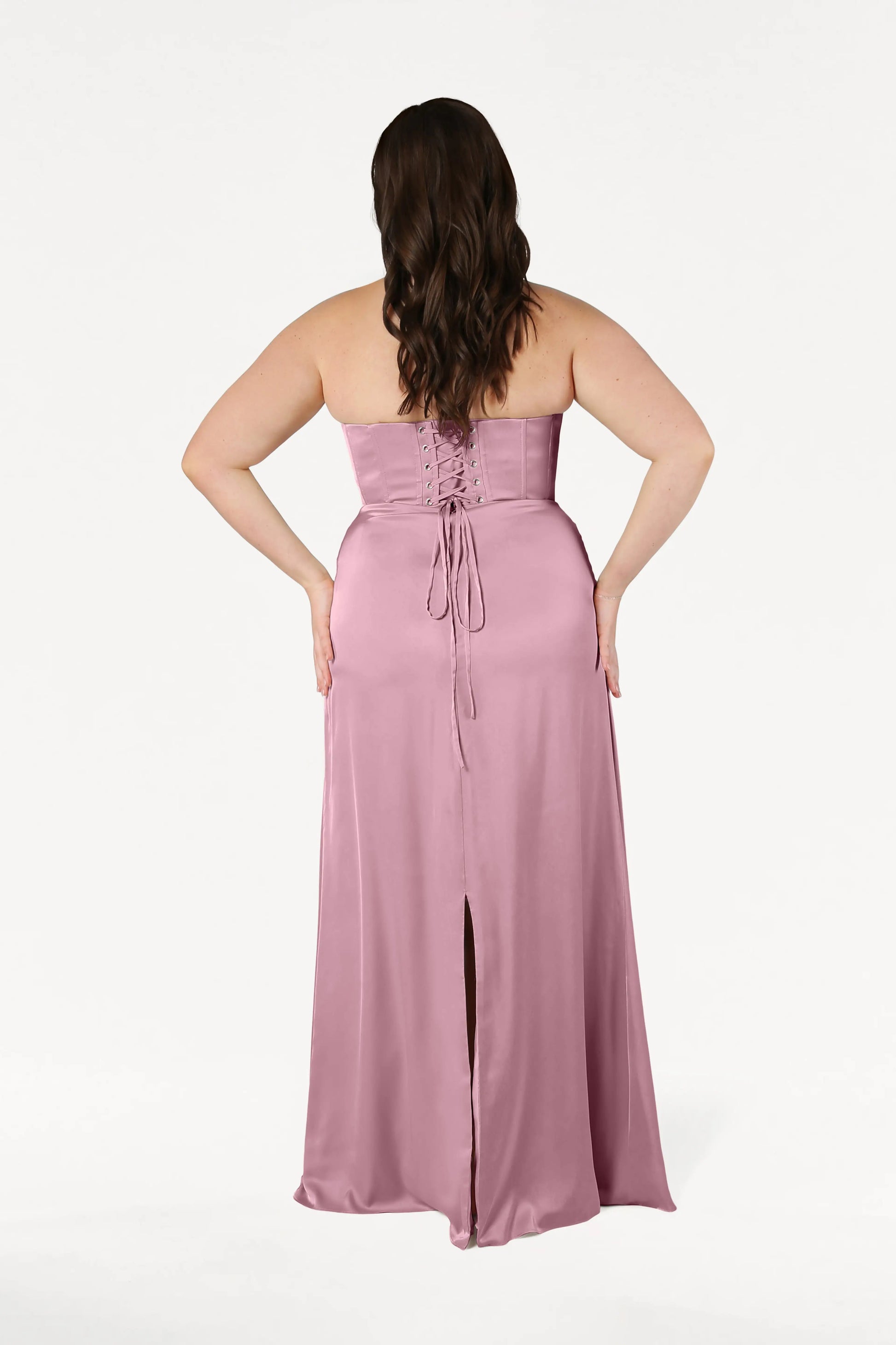 A woman with long brown hair, seen from behind, wears the Selena Bridesmaid Dress in Rose Pink Satin by TH&TH, featuring a lace-up corset bodice and center slit. She stands against a plain white background.