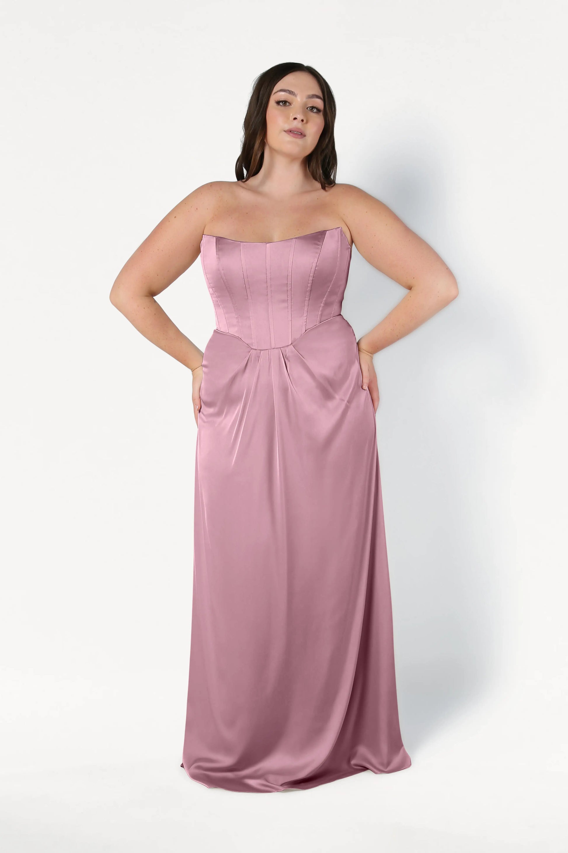 A woman wears the Selena Bridesmaid Dress in Rose Pink Satin by TH&TH, a strapless, floor-length gown with a fitted corset bodice and gathered waist, standing against a plain white background.
