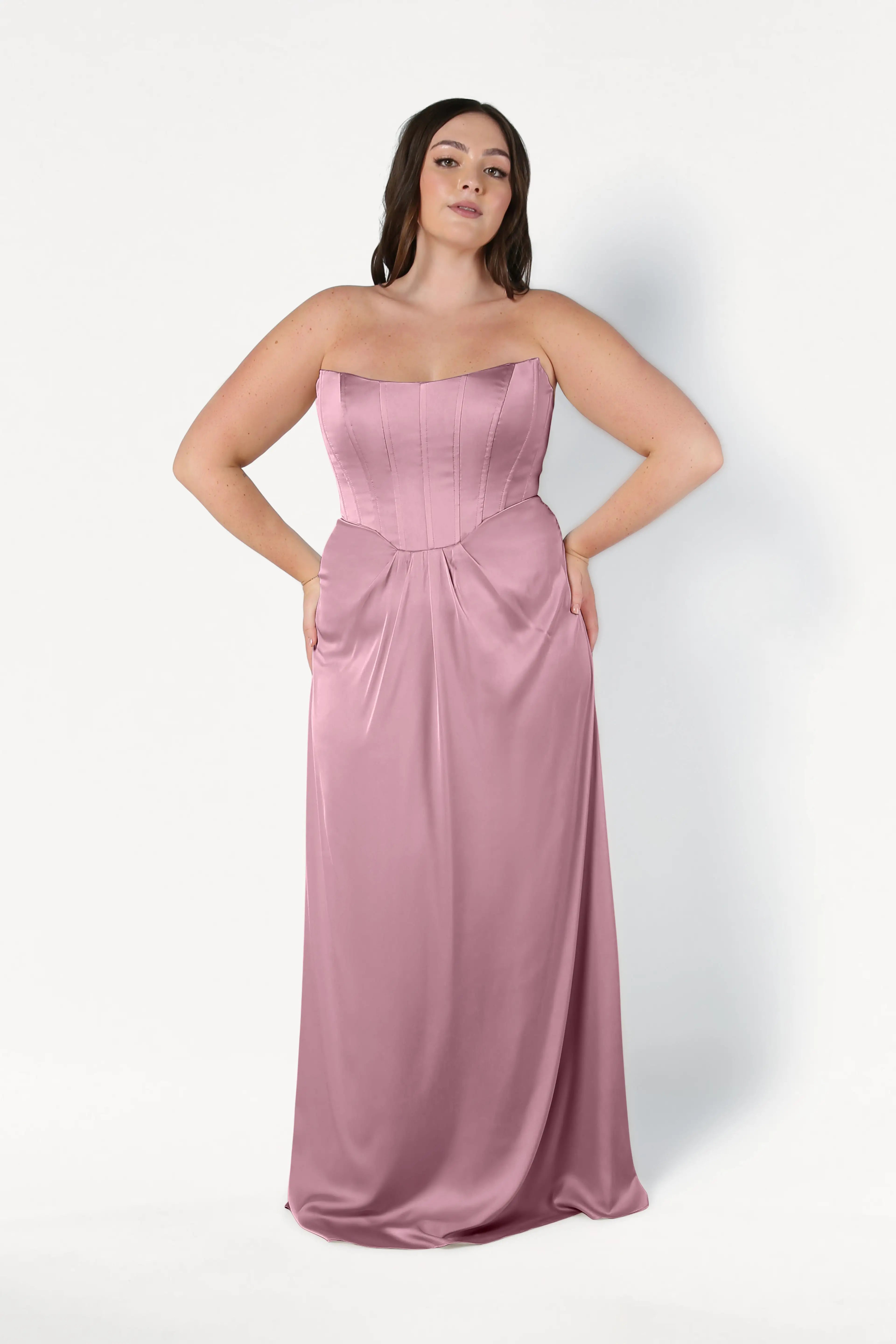A woman wears the Selena Bridesmaid Dress in Rose Pink Satin by TH&TH, a strapless, floor-length gown with a fitted corset bodice and gathered waist, standing against a plain white background.