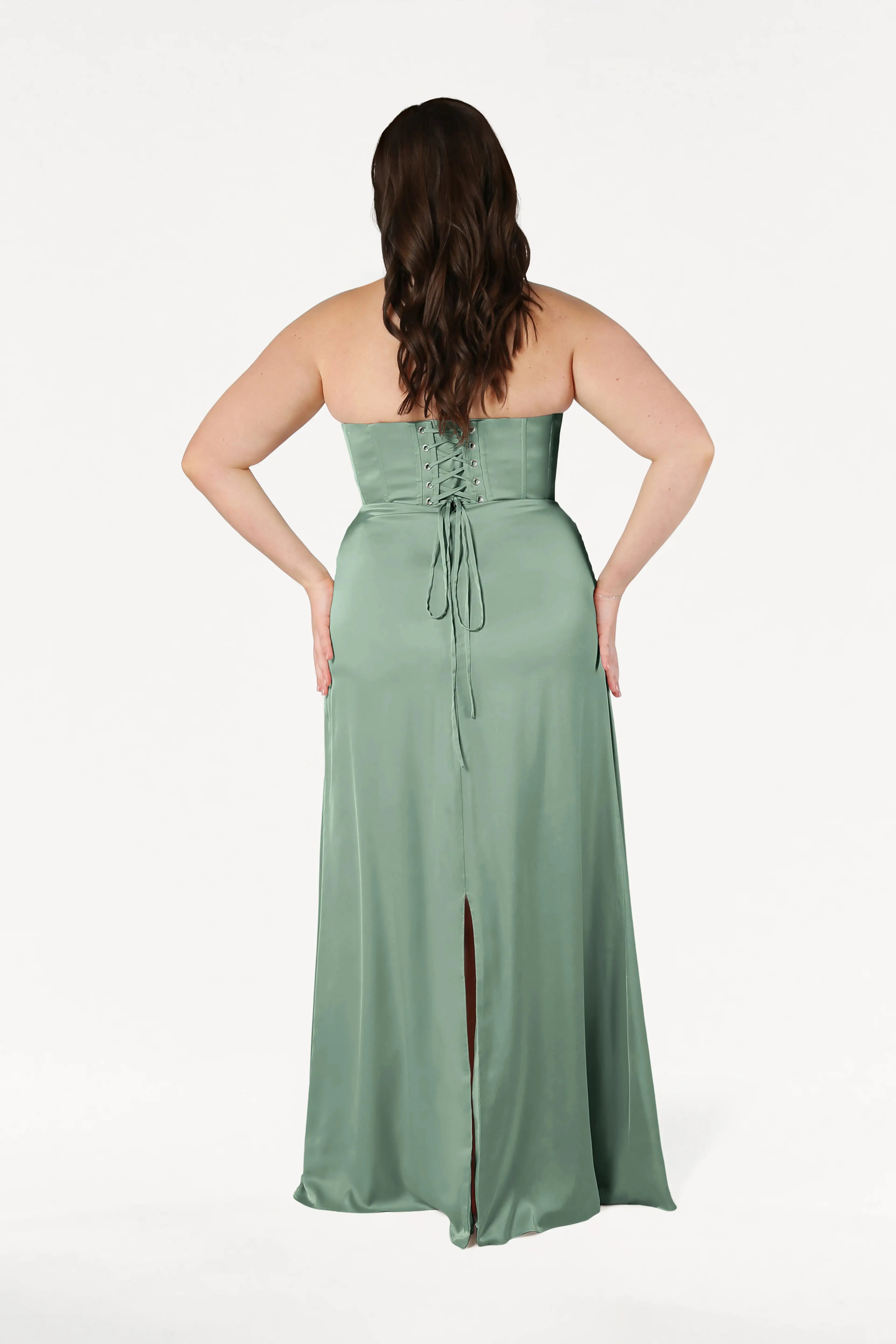 A woman with wavy brown hair models the TH&TH Selena Bridesmaid Dress in sage green satin, featuring a corset bodice, lace-up back, and high slit, standing with her back to the camera against a white background.
