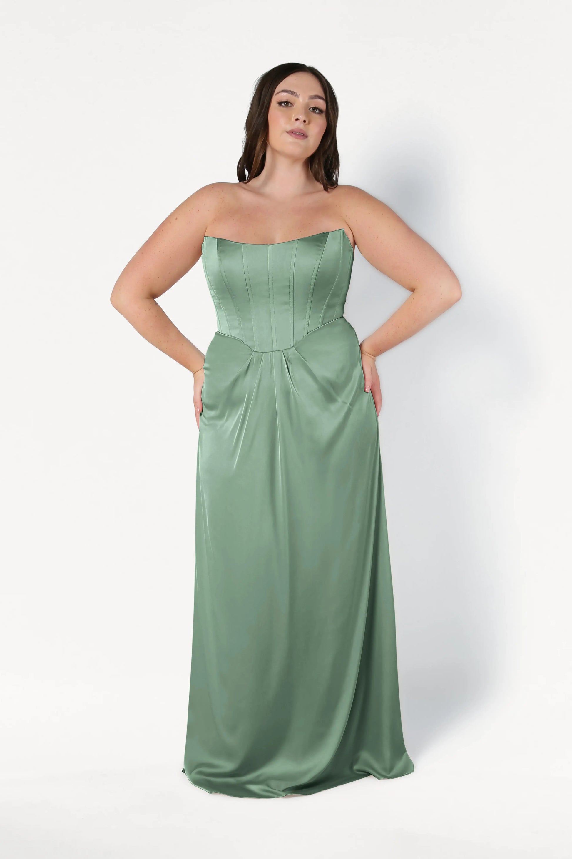 A woman models the TH&TH Selena Bridesmaid Dress in Sage Green Satin, featuring a strapless corset bodice and flowing skirt, as she stands against a plain white background with hands on her hips.