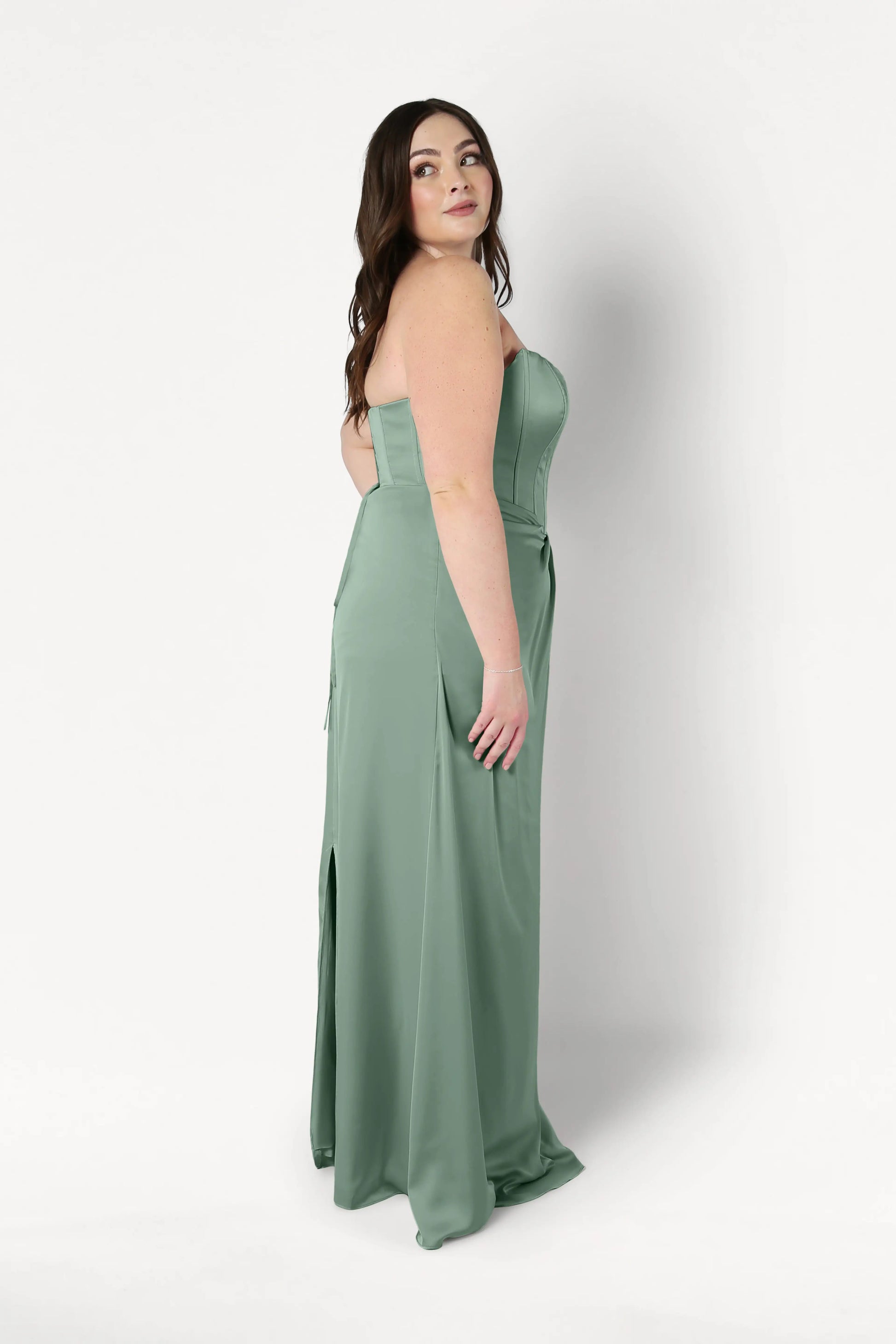 A woman with long brown hair models the TH&TH Selena Bridesmaid Dress in Sage Green Satin—a size-inclusive, strapless gown with a corset bodice—standing sideways and looking over her shoulder against a plain white background.