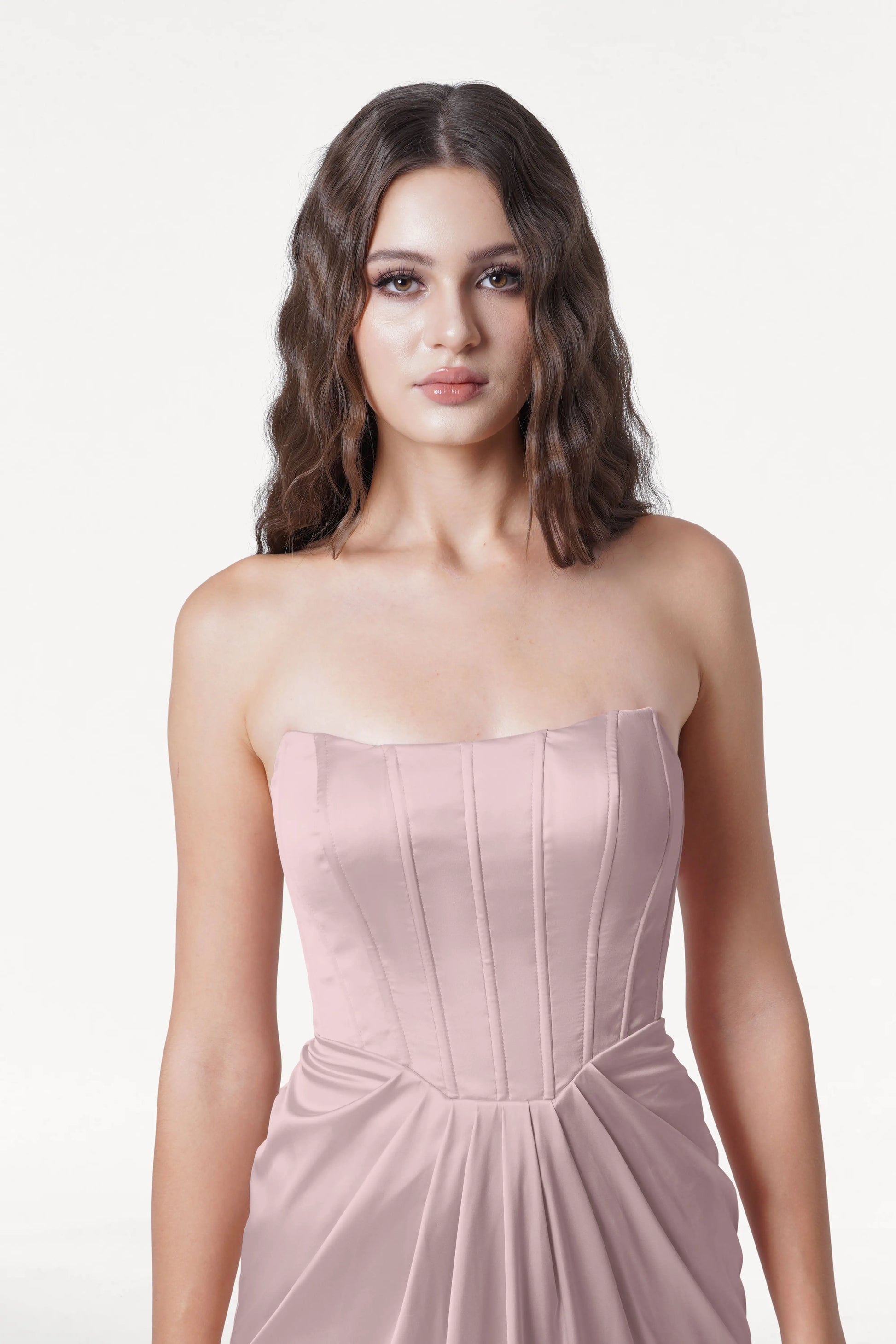 A woman with long, wavy brown hair models the size-inclusive Selena Bridesmaid Dress in Smoked Blush Satin by TH&TH, featuring a fitted corset bodice and draped details, against a plain white background.
