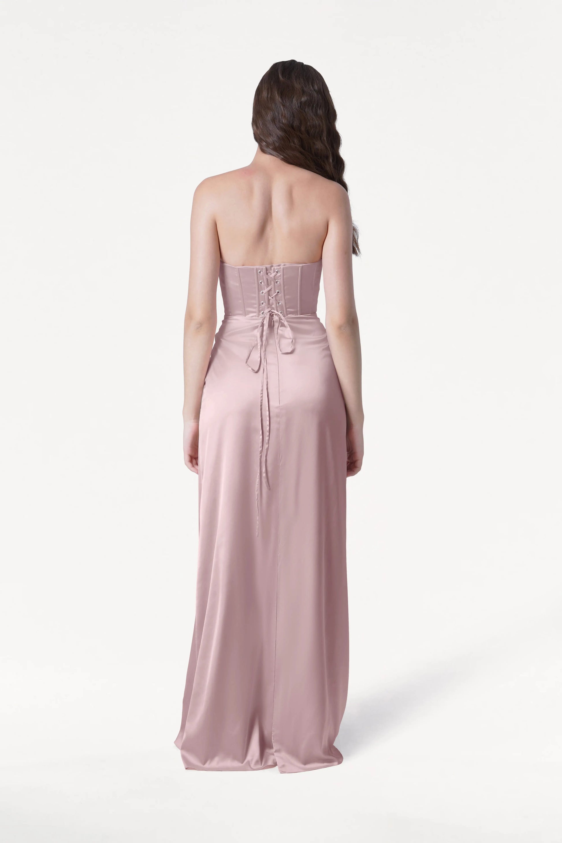 A woman with long brown hair faces away, wearing the TH&TH Selena Bridesmaid Dress in Smoked Blush Satin—a size-inclusive gown featuring a corset bodice and lace-up back—against a plain white background.