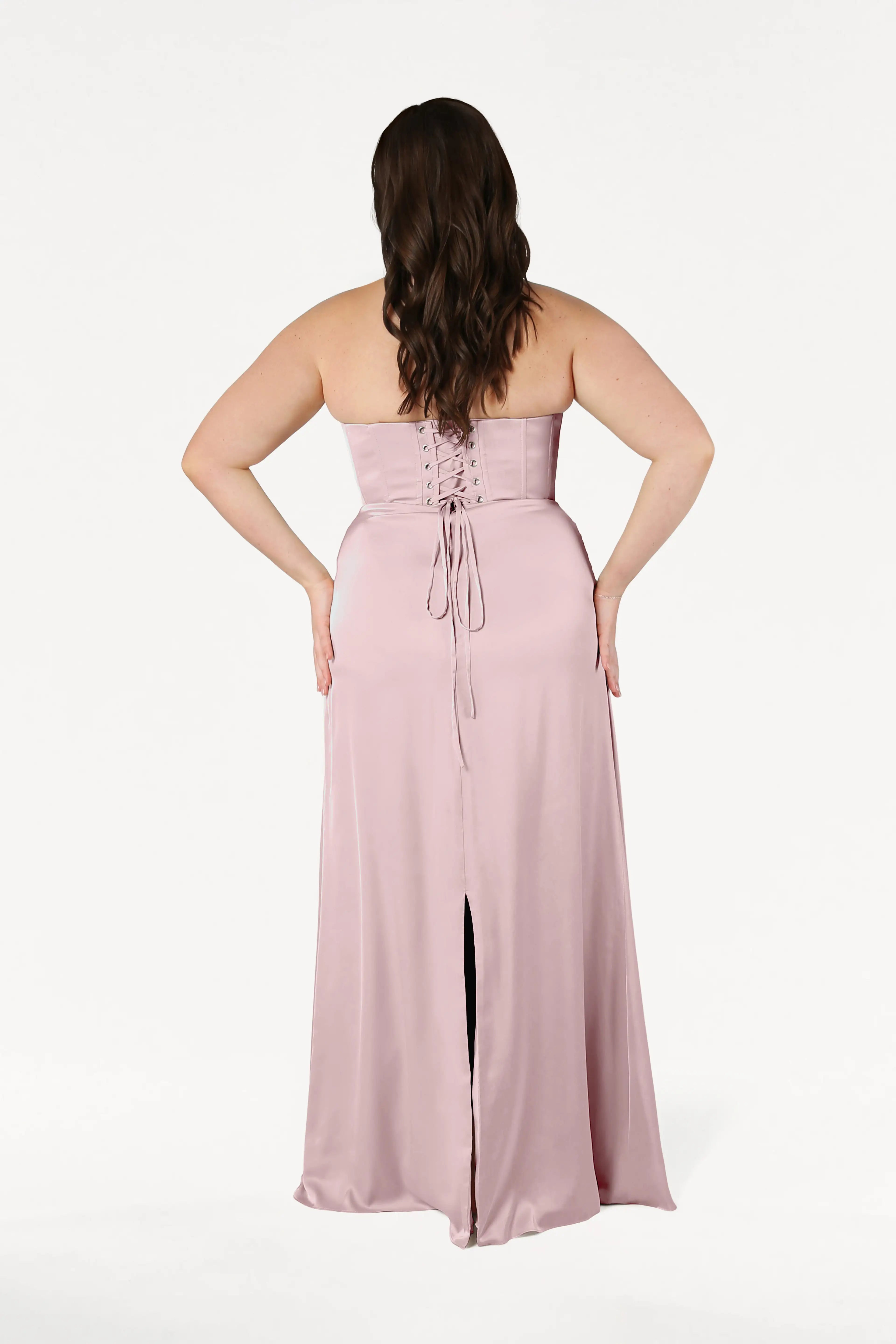 A woman with long brown hair faces away, wearing the Selena Bridesmaid Dress in Smoked Blush Satin by TH&TH—a strapless, floor-length gown with a corset bodice, lace-up back, and center slit. The background is plain white.