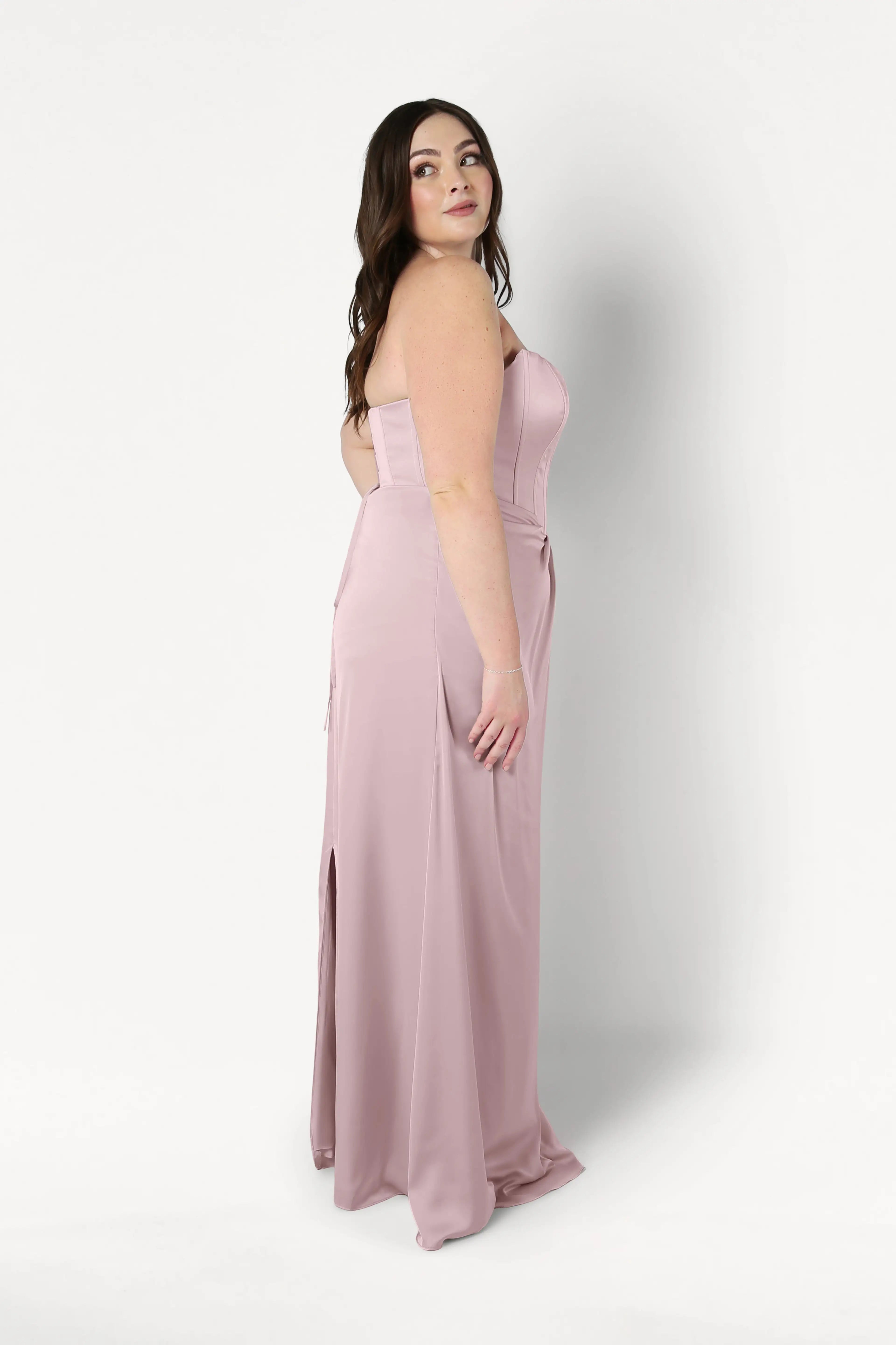 A woman with long brown hair poses in profile against a white background, wearing the TH&TH Selena Bridesmaid Dress in smoked blush satin, featuring a strapless corset bodice and side slit.