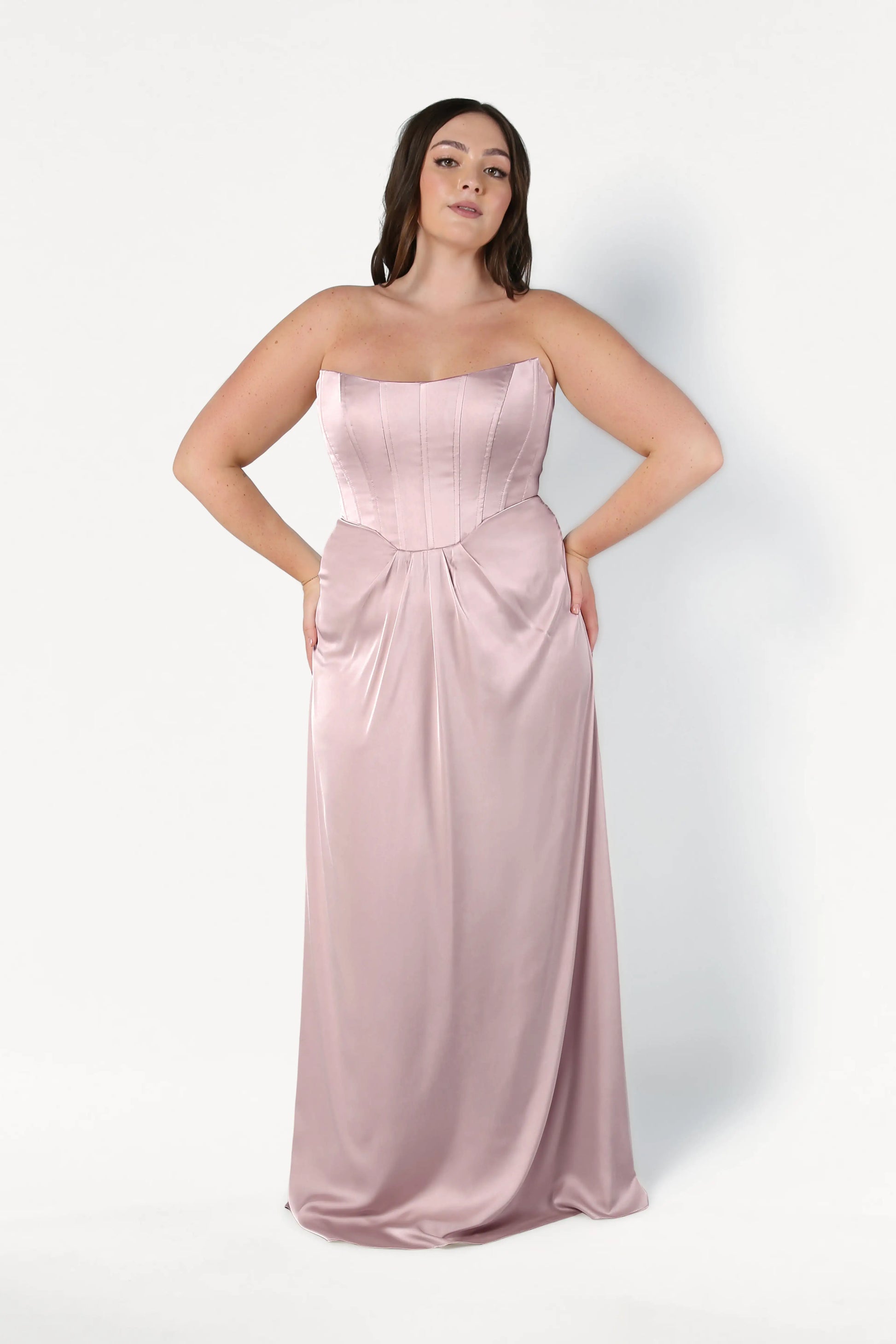 A woman with long dark hair poses against a white background in the Selena Bridesmaid Dress in Smoked Blush Satin by TH&TH, featuring a strapless, floor-length satin silhouette with a fitted corset bodice and flowing skirt. Her hands rest on her hips.