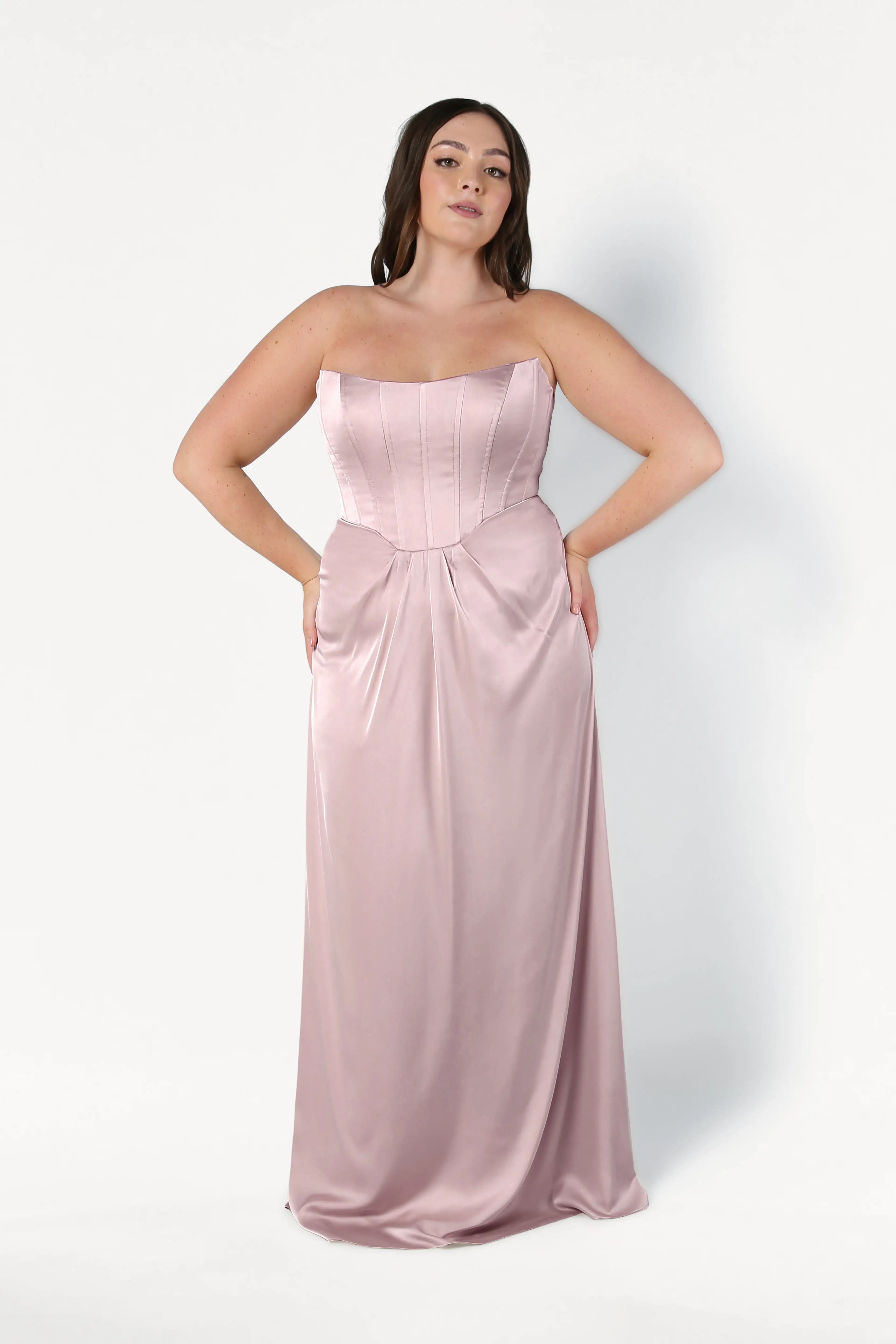 A woman with long dark hair poses against a white background in the Selena Bridesmaid Dress in Smoked Blush Satin by TH&TH, featuring a strapless, floor-length satin silhouette with a fitted corset bodice and flowing skirt. Her hands rest on her hips.
