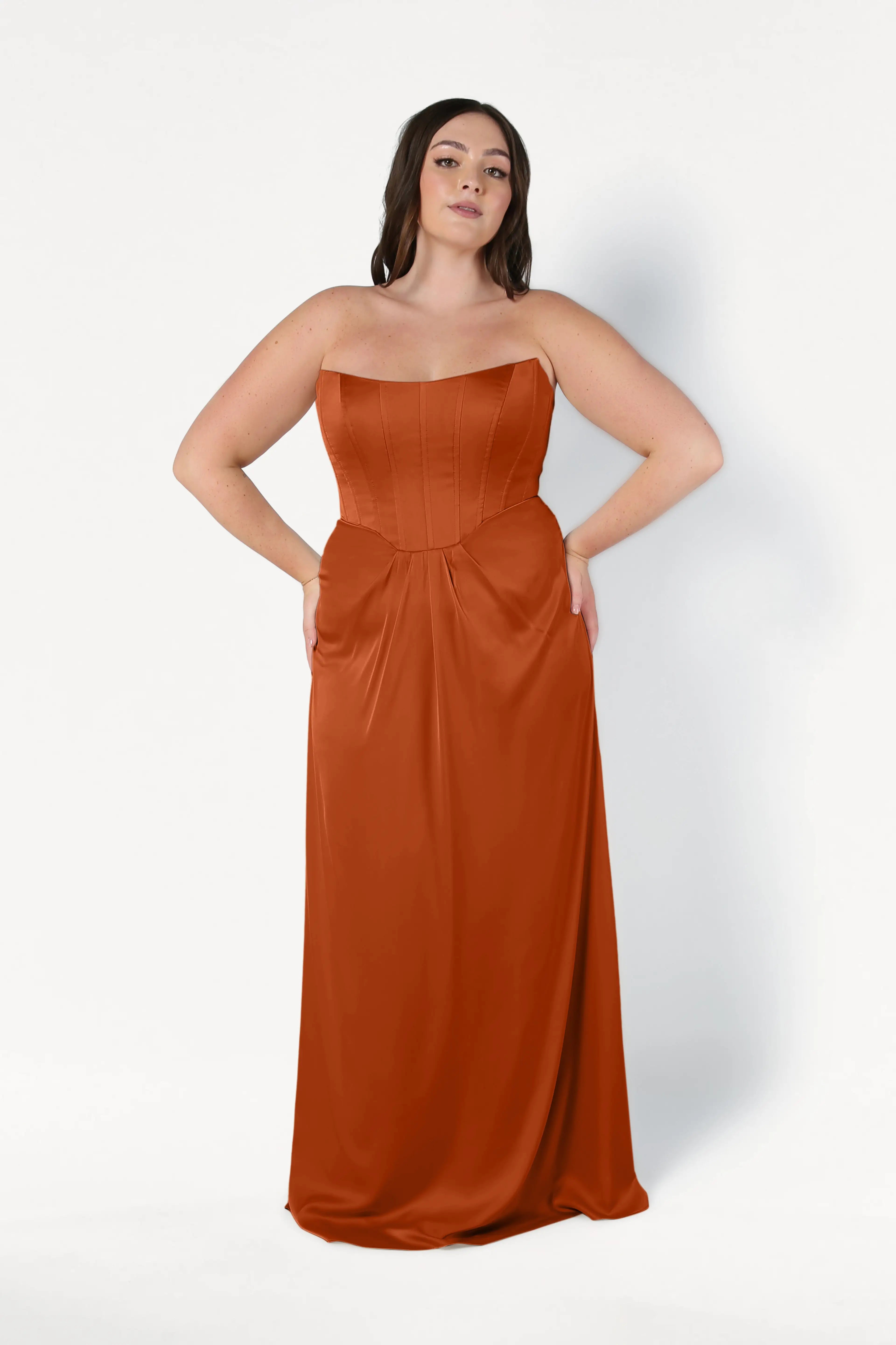 A woman models the Selena Bridesmaid Dress in Terracotta Satin by TH&TH, a size-inclusive, floor-length gown with a strapless corset bodice and pockets, standing confidently against a white background.