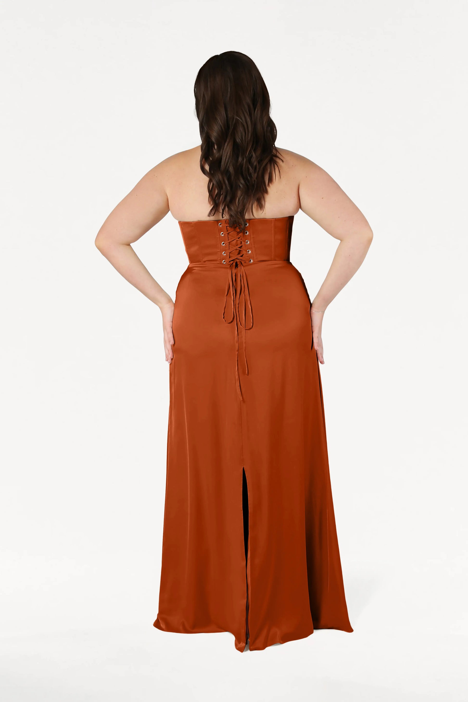 A woman with long brown hair stands with her back to the camera, wearing the size-inclusive TH&TH Selena Bridesmaid Dress in Terracotta Satin, featuring a corset bodice, lace-up back, and front slit. The background is plain white.