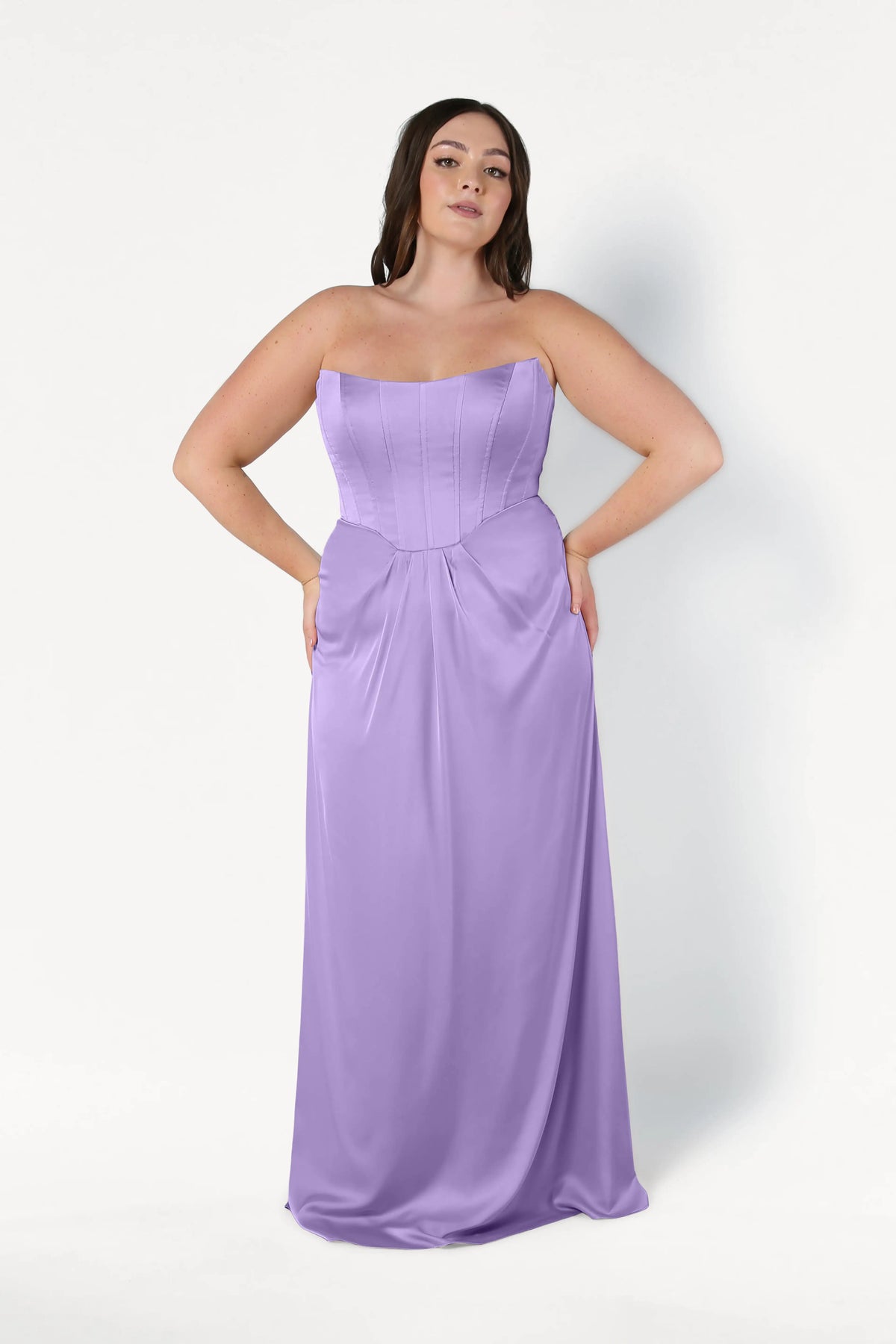 A woman poses against a white background in the Selena Bridesmaid Dress in Violet Satin by TH&TH, featuring a strapless, floor-length design with a fitted corset bodice, flowing skirt, and size-inclusive fit.