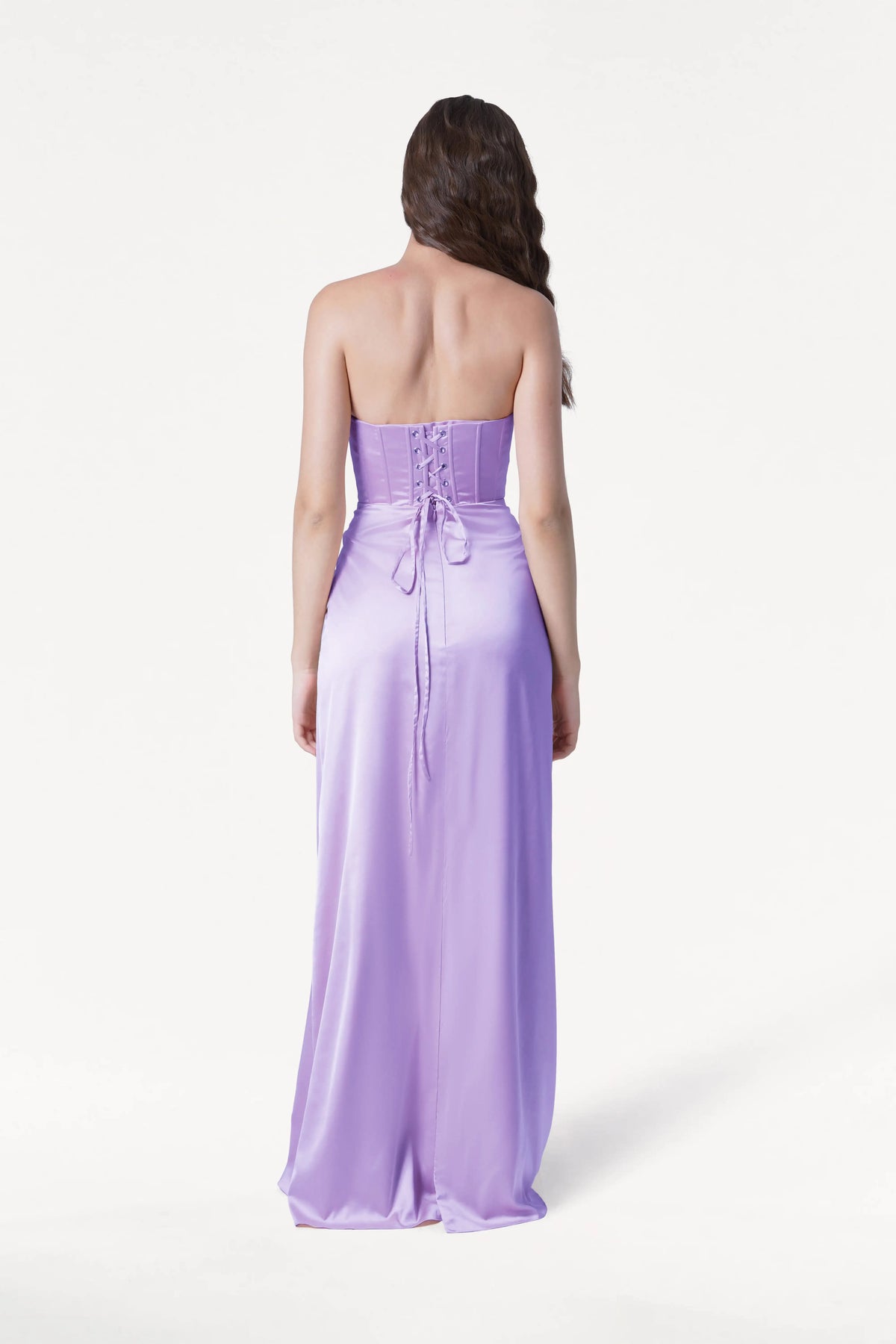 A woman, seen from behind, wears the TH&TH Selena Bridesmaid Dress in Violet Satin—a size-inclusive gown featuring a lace-up corset bodice and floor-length skirt—standing against a plain white background.