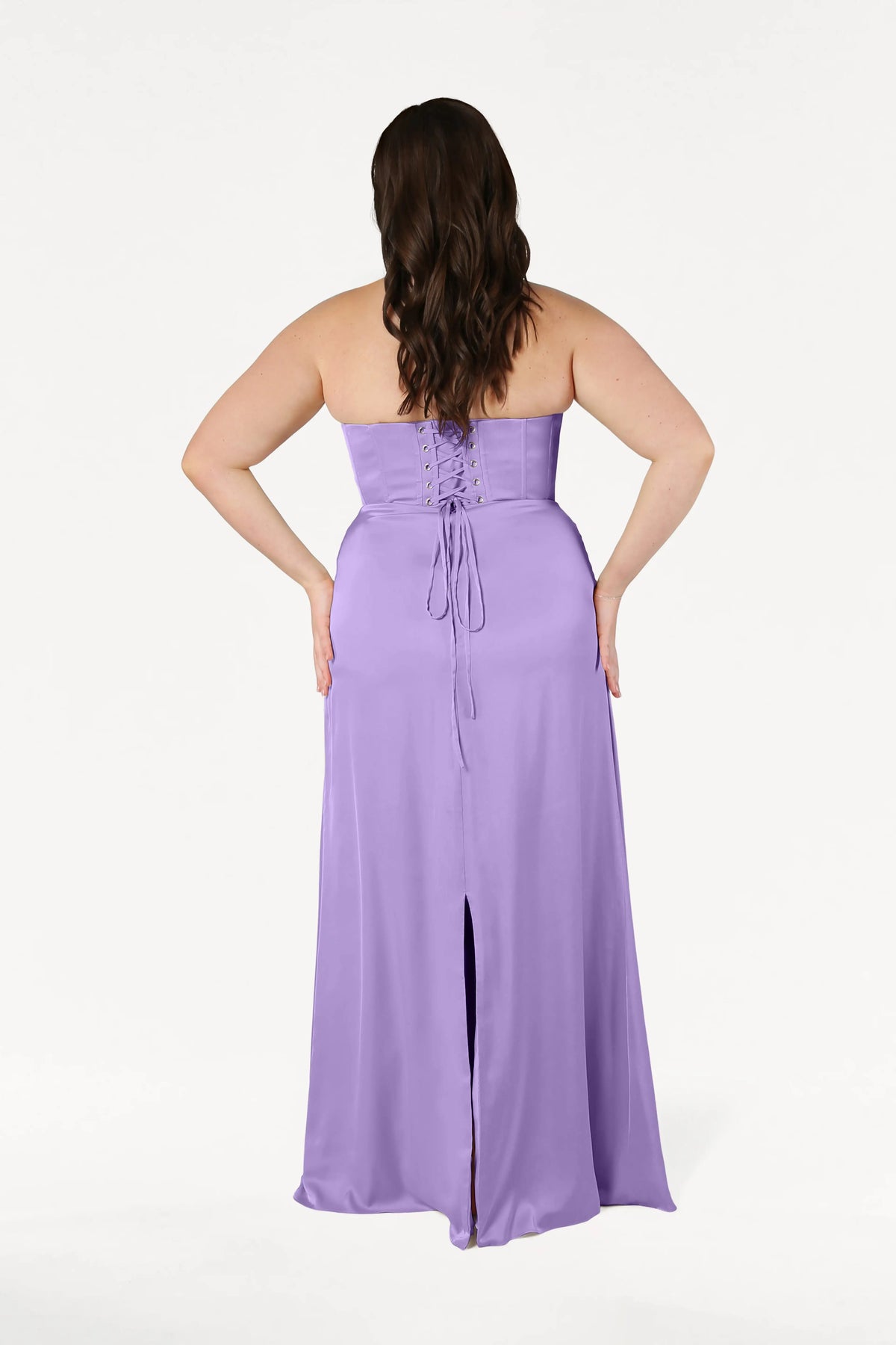 A woman models the TH&TH Selena Bridesmaid Dress in Violet Satin, a size-inclusive, strapless gown with a corset bodice, lace-up back, and center slit, standing hands on hips against a plain white background.