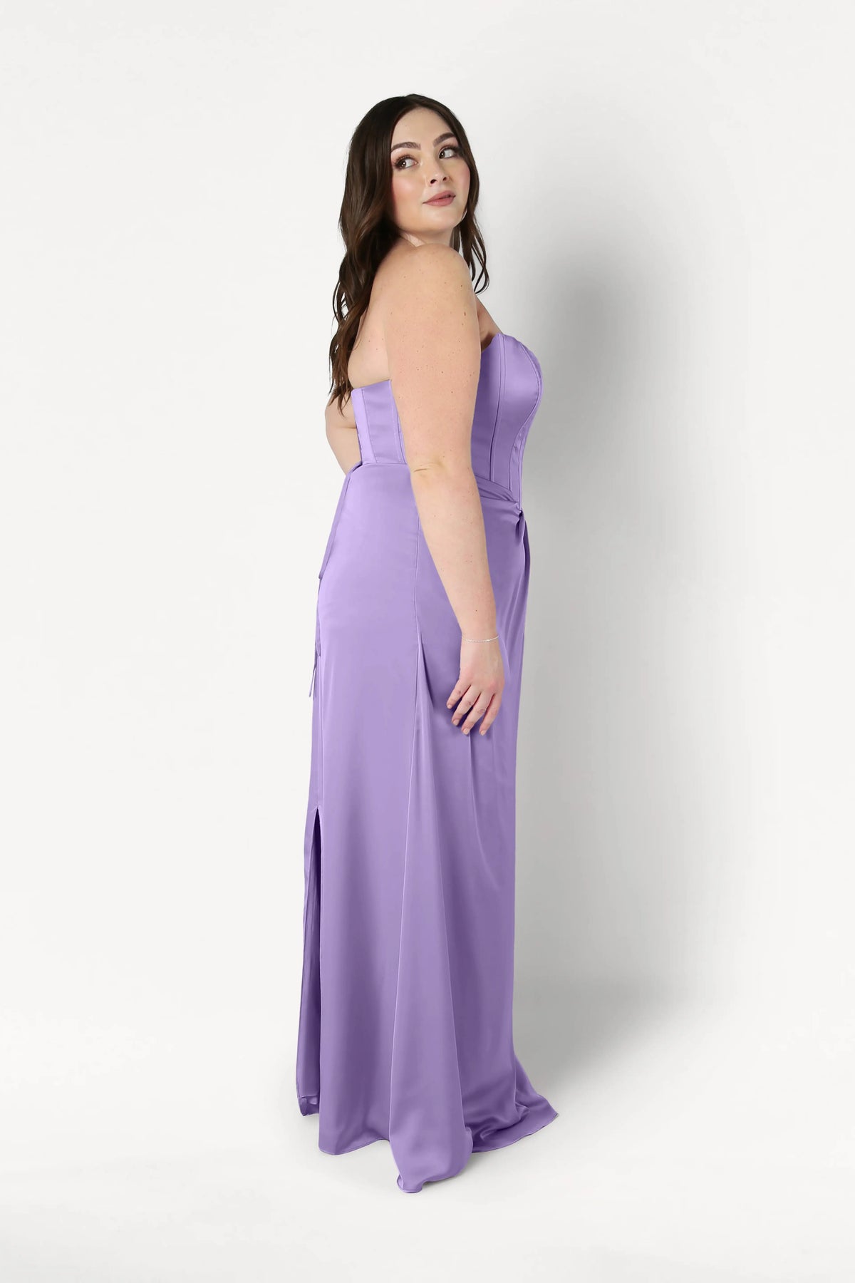 A woman with long brown hair wears the TH&TH Selena Bridesmaid Dress in Violet Satin, featuring a strapless corset bodice and floor-length silhouette, standing sideways and looking over her shoulder against a plain white background.