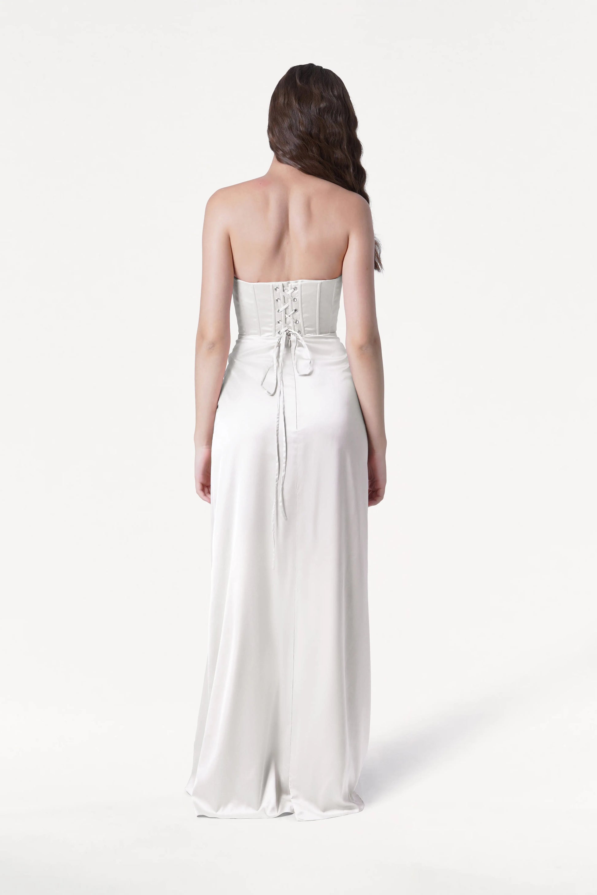A woman with long brown hair faces away, wearing the TH&TH Selena Dress in Ivory Satin—a strapless gown with a corset bodice, lace-up back, and flowing satin skirt—against a plain white background.