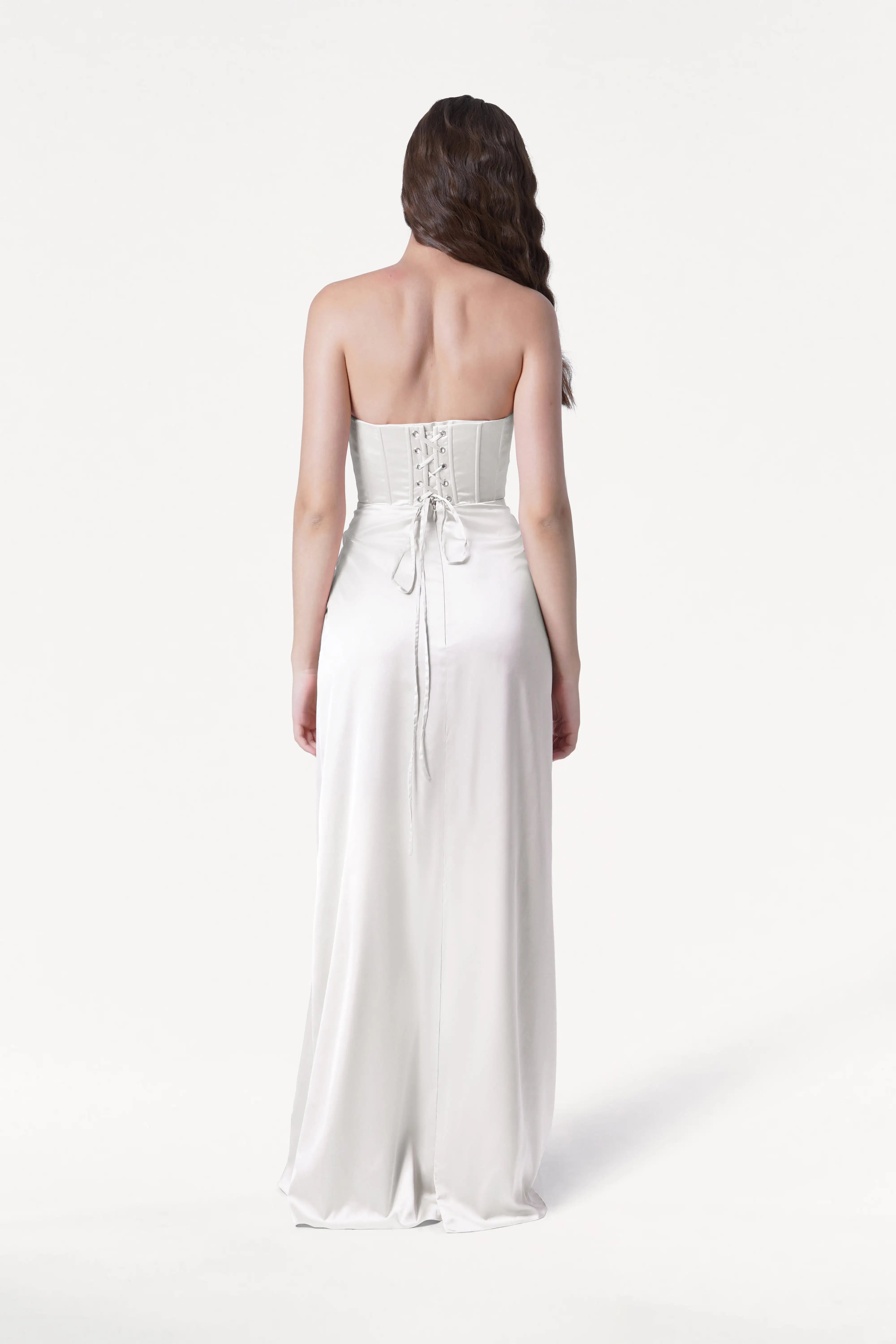 A woman with long brown hair faces away, wearing the TH&TH Selena Dress in Ivory Satin—a strapless gown with a corset bodice, lace-up back, and flowing satin skirt—against a plain white background.