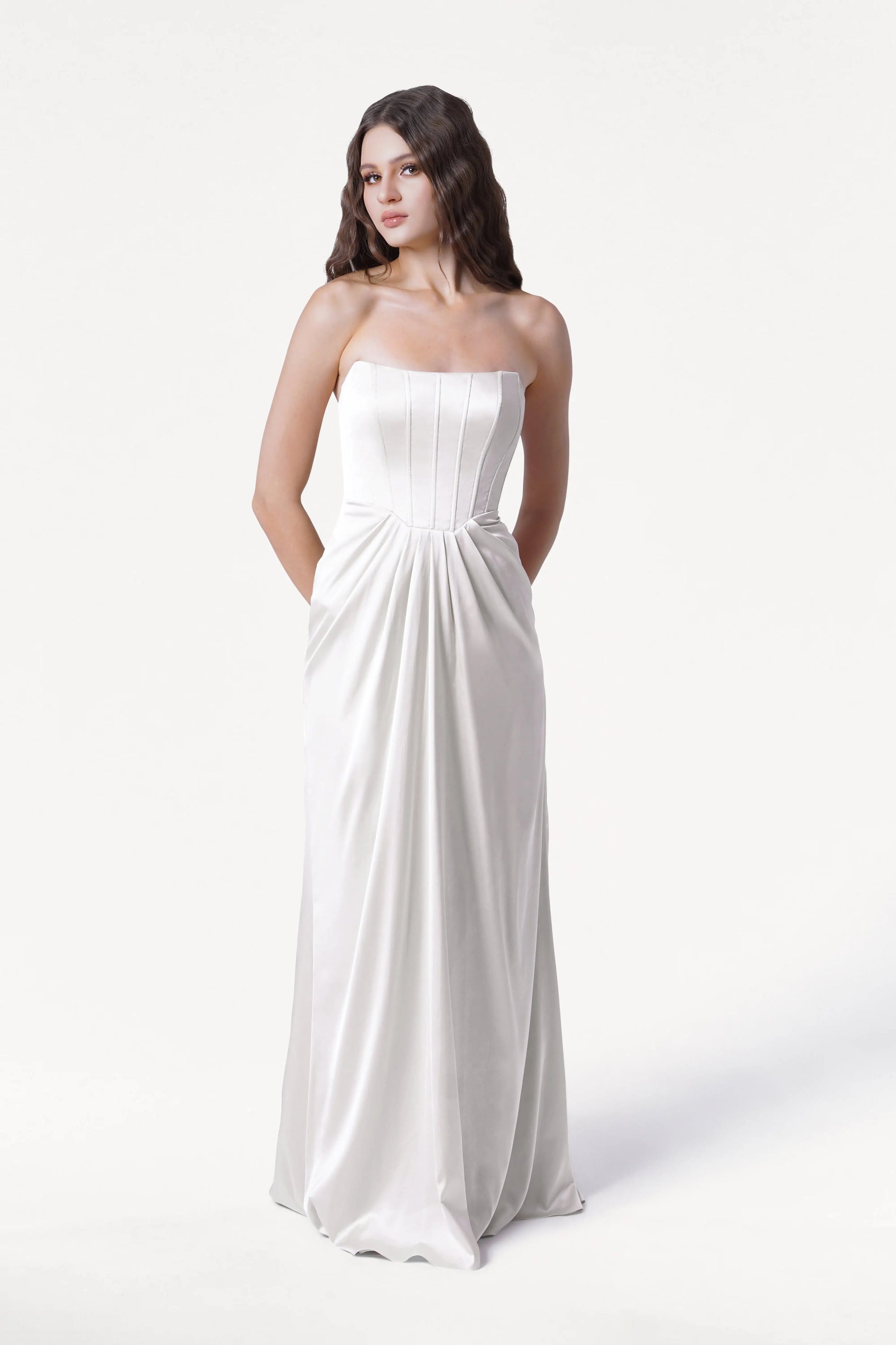A woman with long, wavy brown hair models the Selena Dress in Ivory Satin by TH&TH—a size-inclusive, strapless gown featuring a pleated corset bodice and pockets—standing against a plain white background.