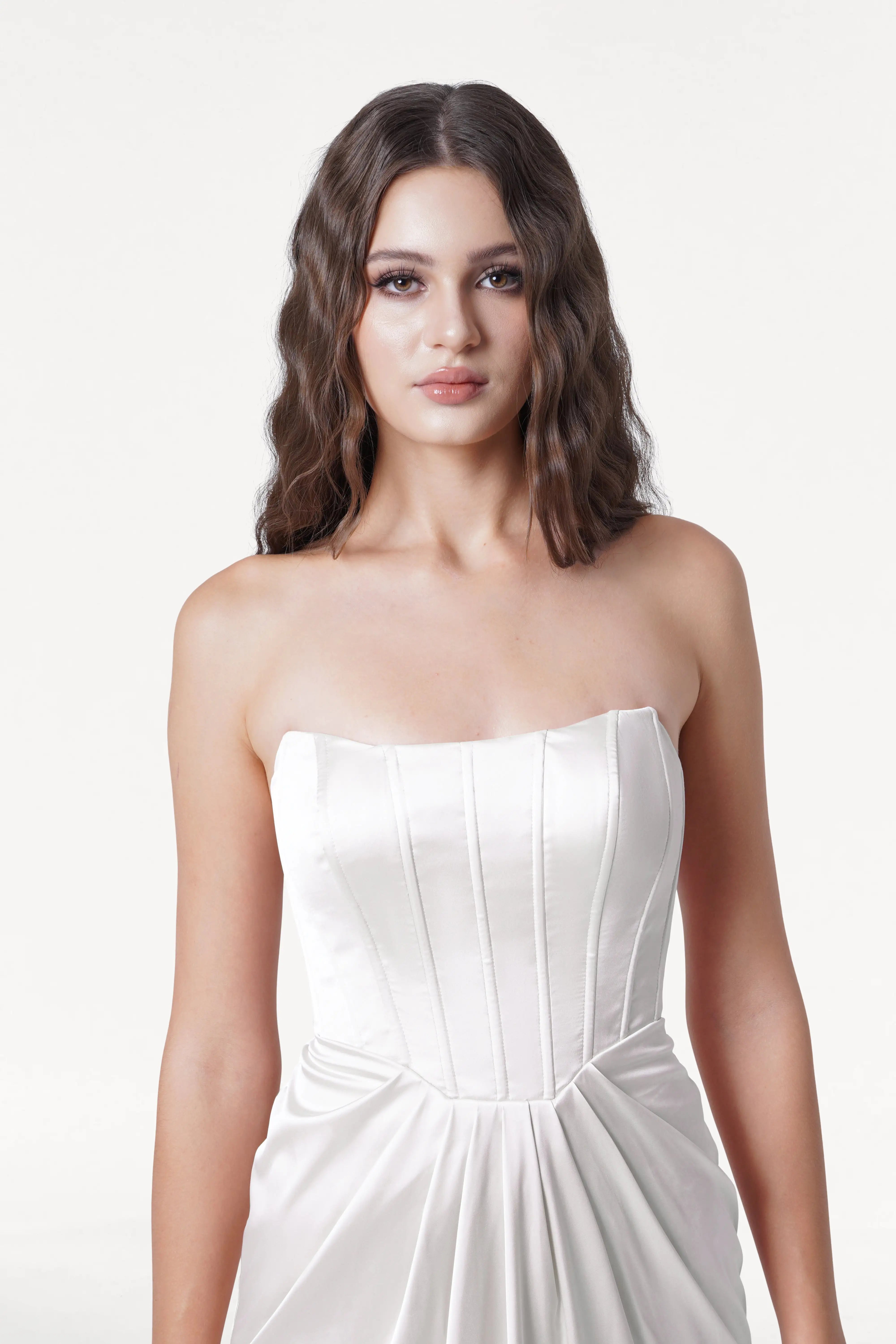 A young woman with long, wavy brown hair wears the TH&TH Selena Dress in Ivory Satin, a strapless satin bridesmaid dress with a corset bodice, standing against a plain white background and gazing at the camera with a neutral expression.