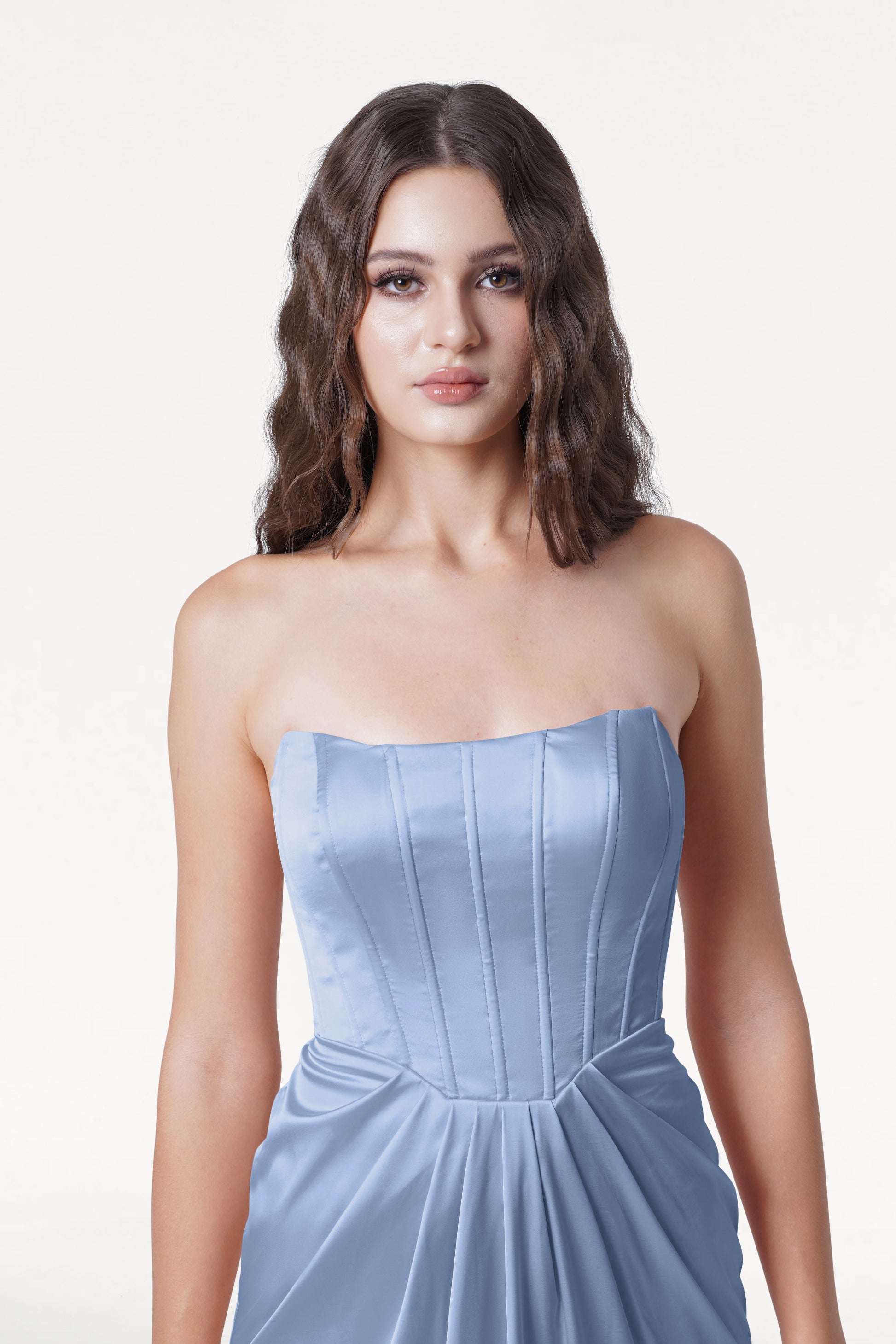 A woman with long, wavy brown hair wears the TH&TH Selena Bridesmaid Dress in Dusty Blue Satin, featuring vertical seams and a corset bodice at the waist, standing against a plain white background.