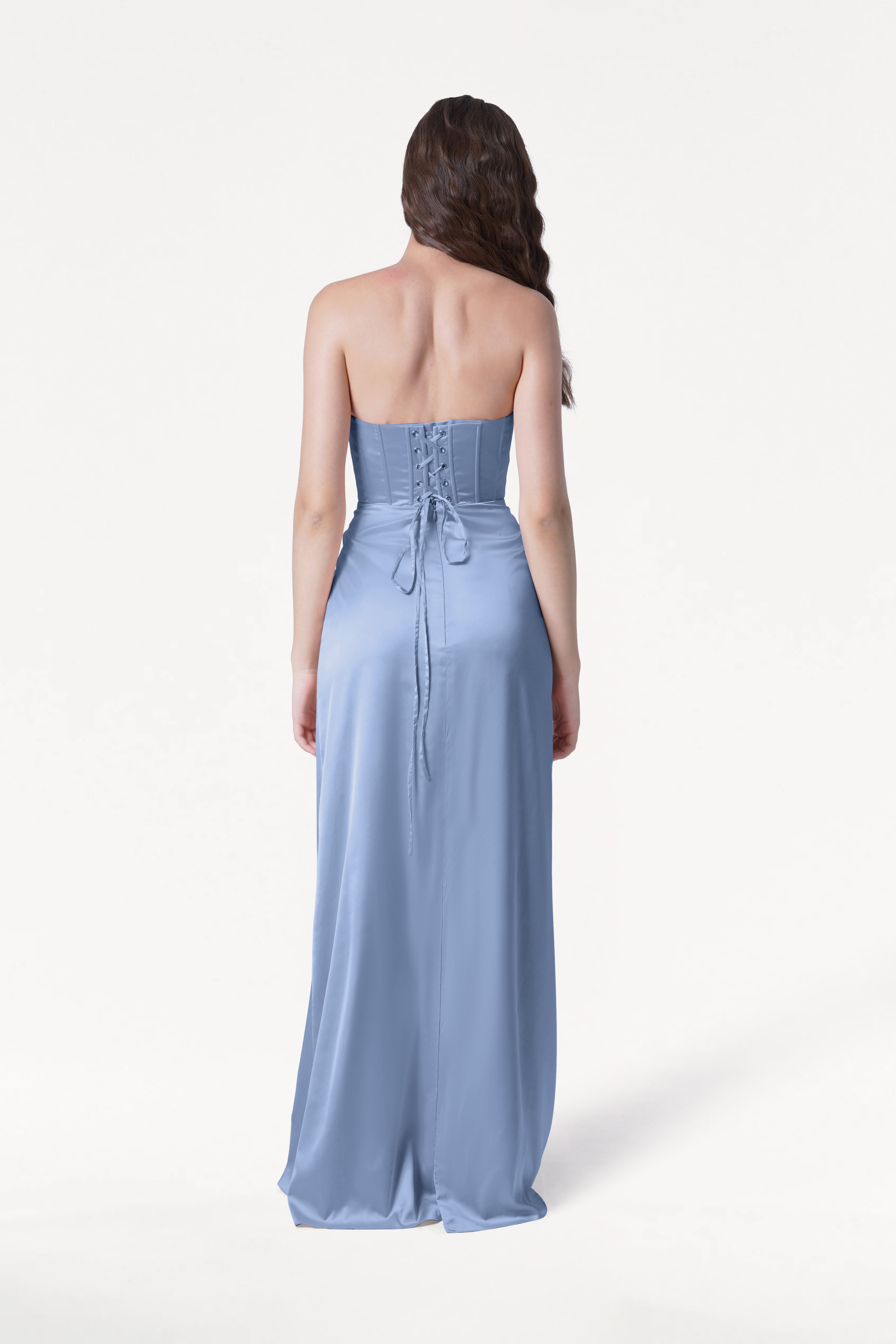 A woman with long brown hair stands with her back to the camera against a white background, wearing the TH&TH Selena Bridesmaid Dress in Dusty Blue Satin—a strapless, floor-length gown featuring a corset bodice.