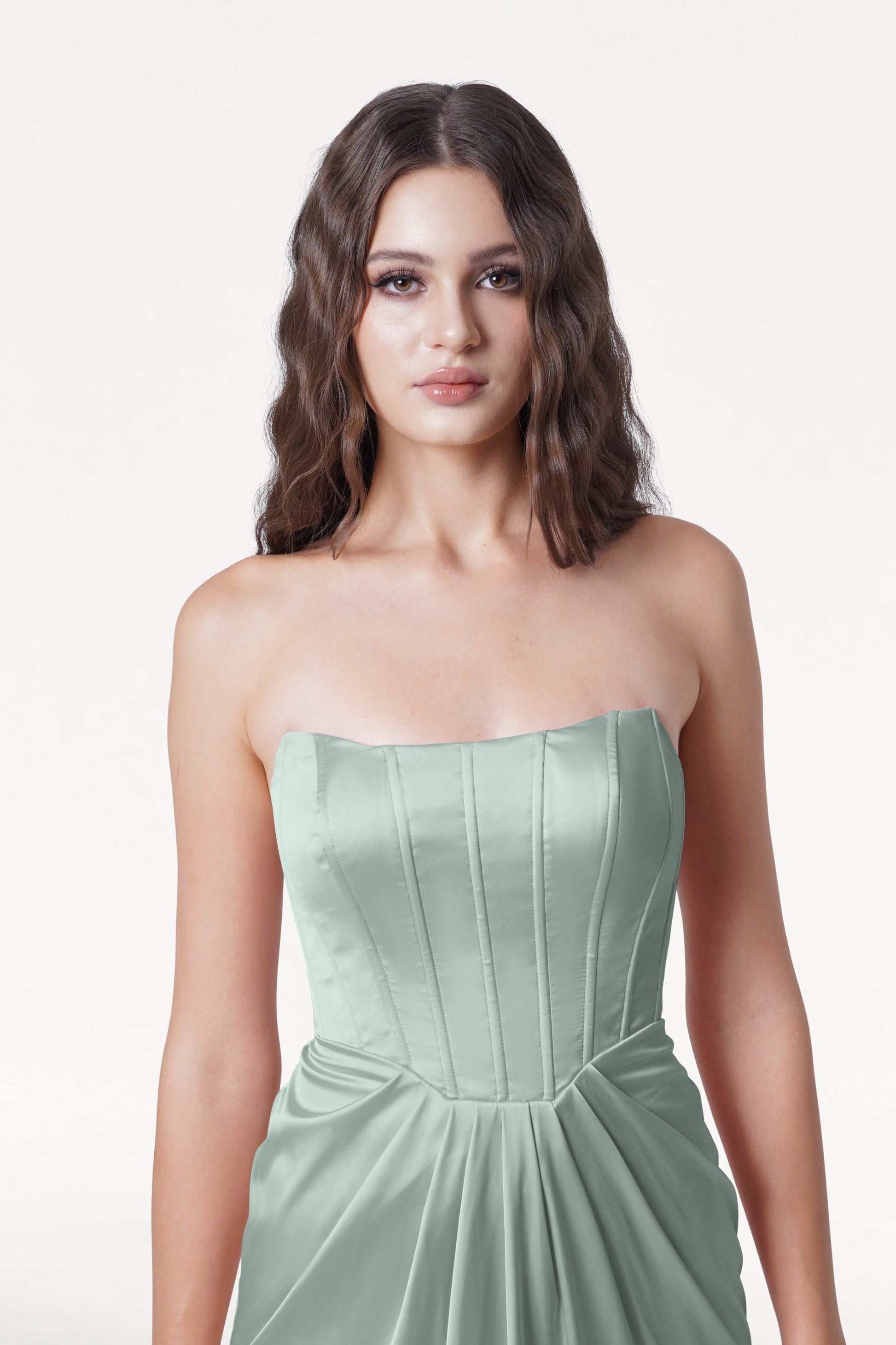 A woman with wavy brown hair wears the TH&TH Selena Bridesmaid Dress in Dusty Eucalyptus Satin, featuring a strapless corset bodice, standing against a plain white background and looking at the camera with a neutral expression.