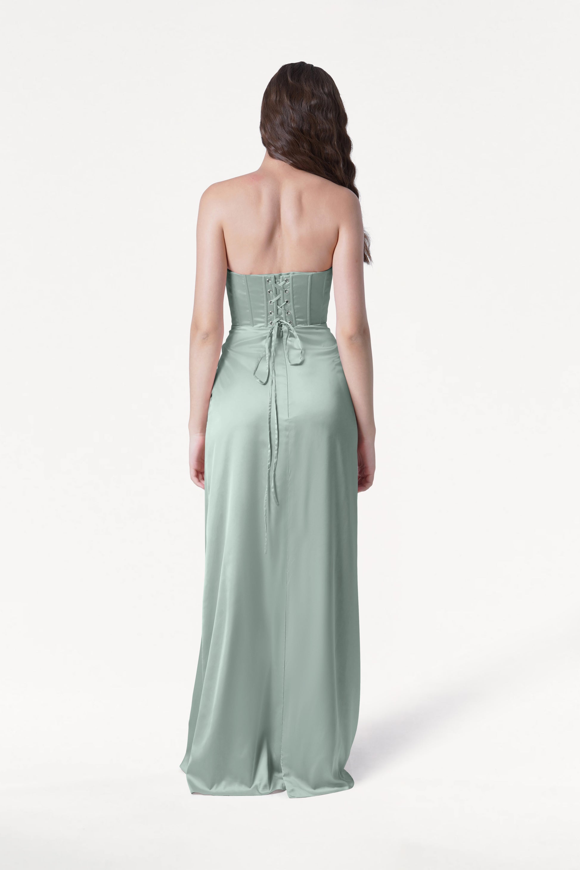 A woman faces away, wearing the TH&TH Selena Bridesmaid Dress in Dusty Eucalyptus Satin. This size-inclusive gown features a corset bodice, lace-up back, and long flowing skirt. Her wavy brown hair drapes over one shoulder against a white background.