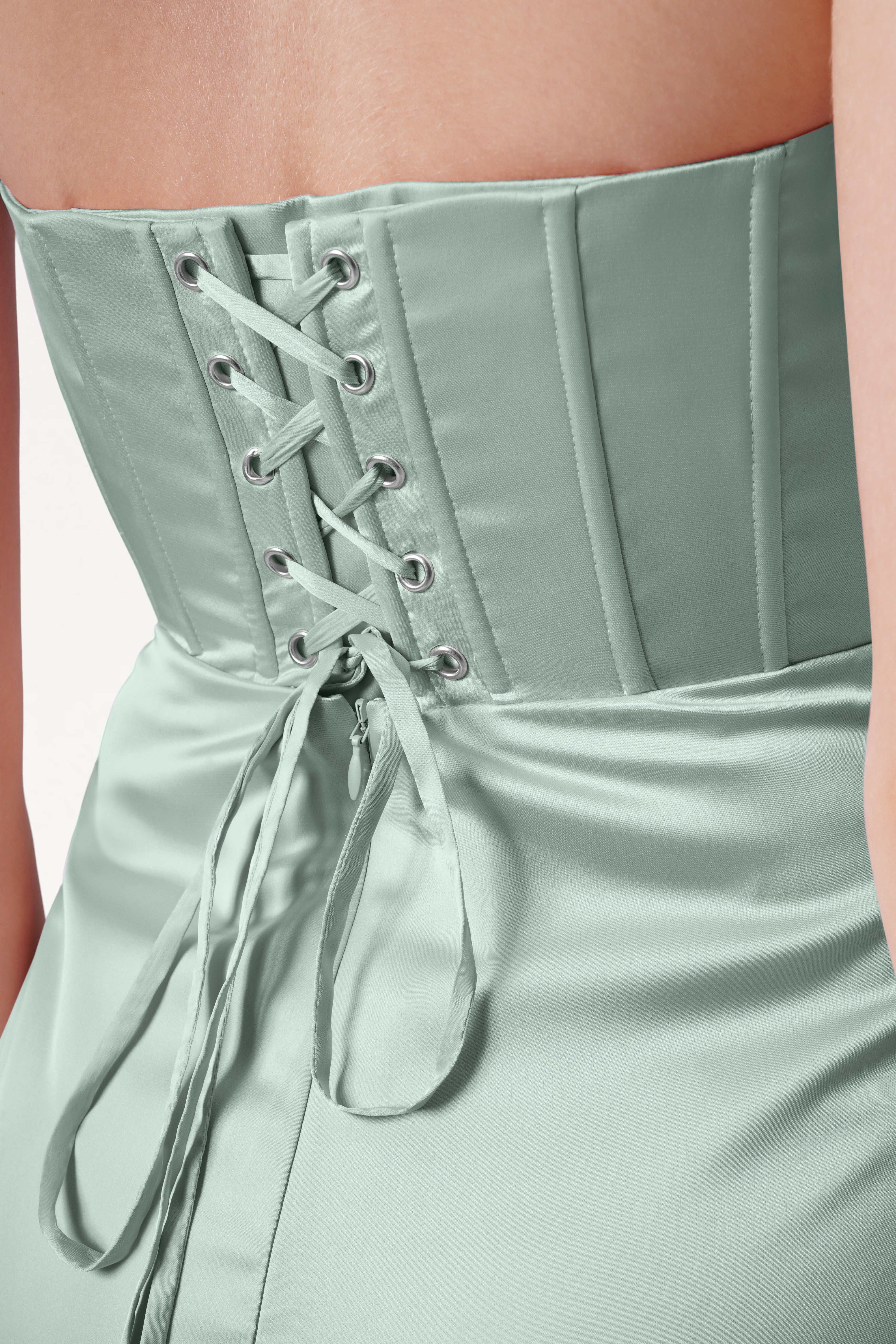A woman wears the Selena Bridesmaid Dress in Dusty Eucalyptus Satin by TH&TH, a size-inclusive gown with a corset bodice, vertical seams, lace-up ribbon back, and smooth, shiny light green fabric.