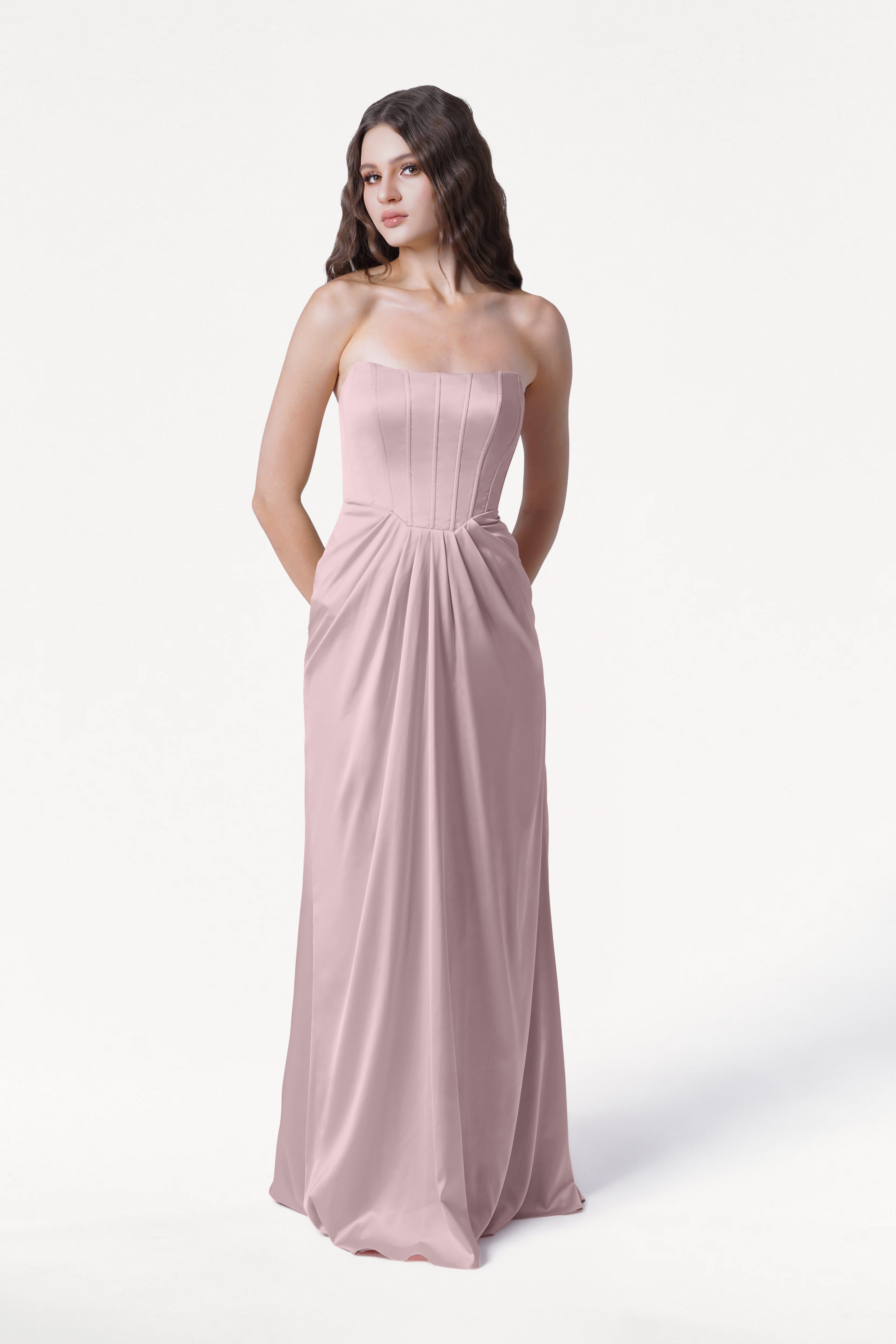 A woman with long brown hair wears the Selena Bridesmaid Dress in Smoked Blush Satin by TH&TH—a size-inclusive, strapless, floor-length gown with a corset bodice and pockets—standing against a plain white background.