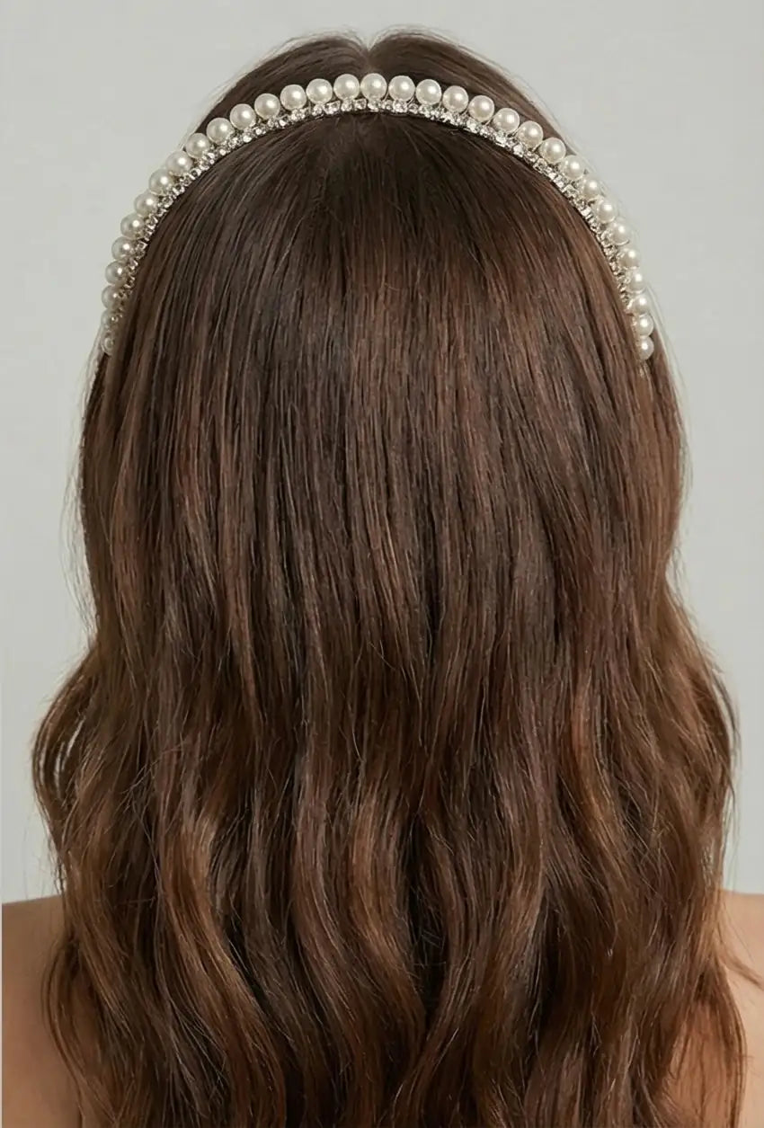 A woman with long, wavy brown hair is shown from the back, wearing the TH&TH Seraphine Crystal Headband. This elegant pearl and crystal bridal accessory rests on her crown, adding a sophisticated touch to her wedding hairstyle.