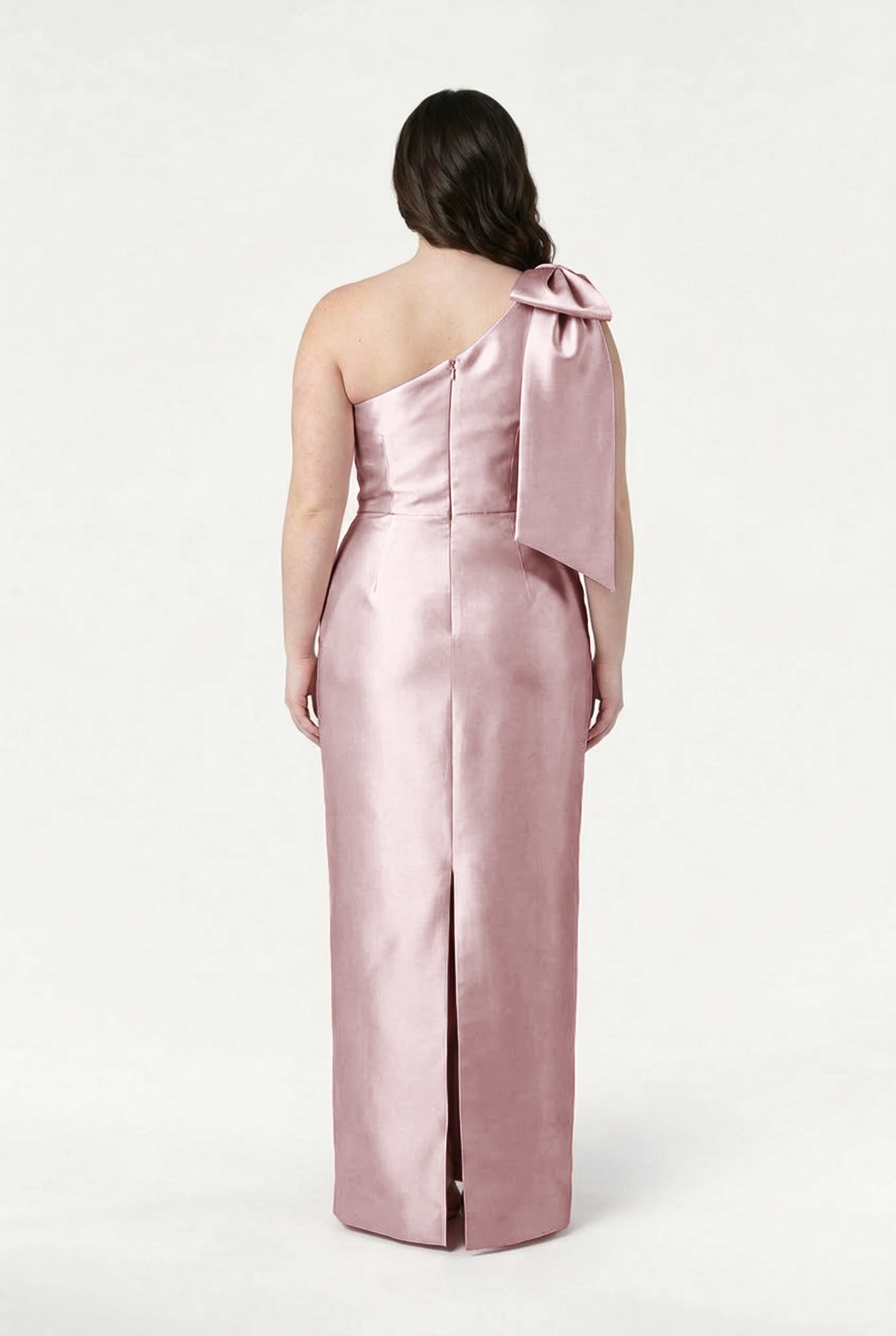 A woman faces away, wearing the TH&TH Ritz Bridesmaid Dress in Smoked Blush Satin Twill—a shiny, floor-length one-shoulder gown with a statement shoulder bow, back slit, and fitted silhouette against a plain white background.