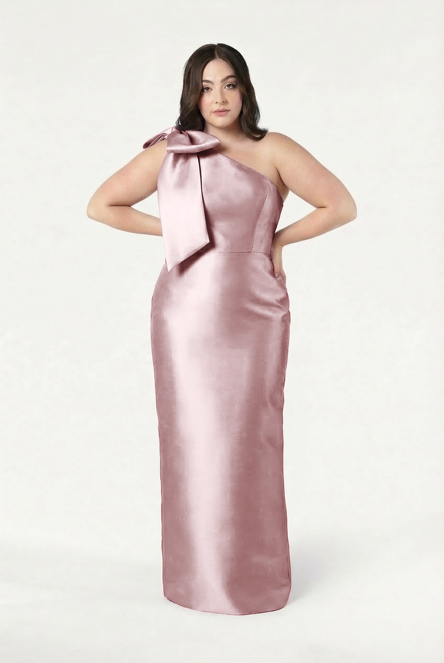 A woman stands hands on hips against a white background, wearing the TH&TH Ritz Bridesmaid Dress in smoked blush satin twill, featuring a long one-shoulder design with a statement bow.