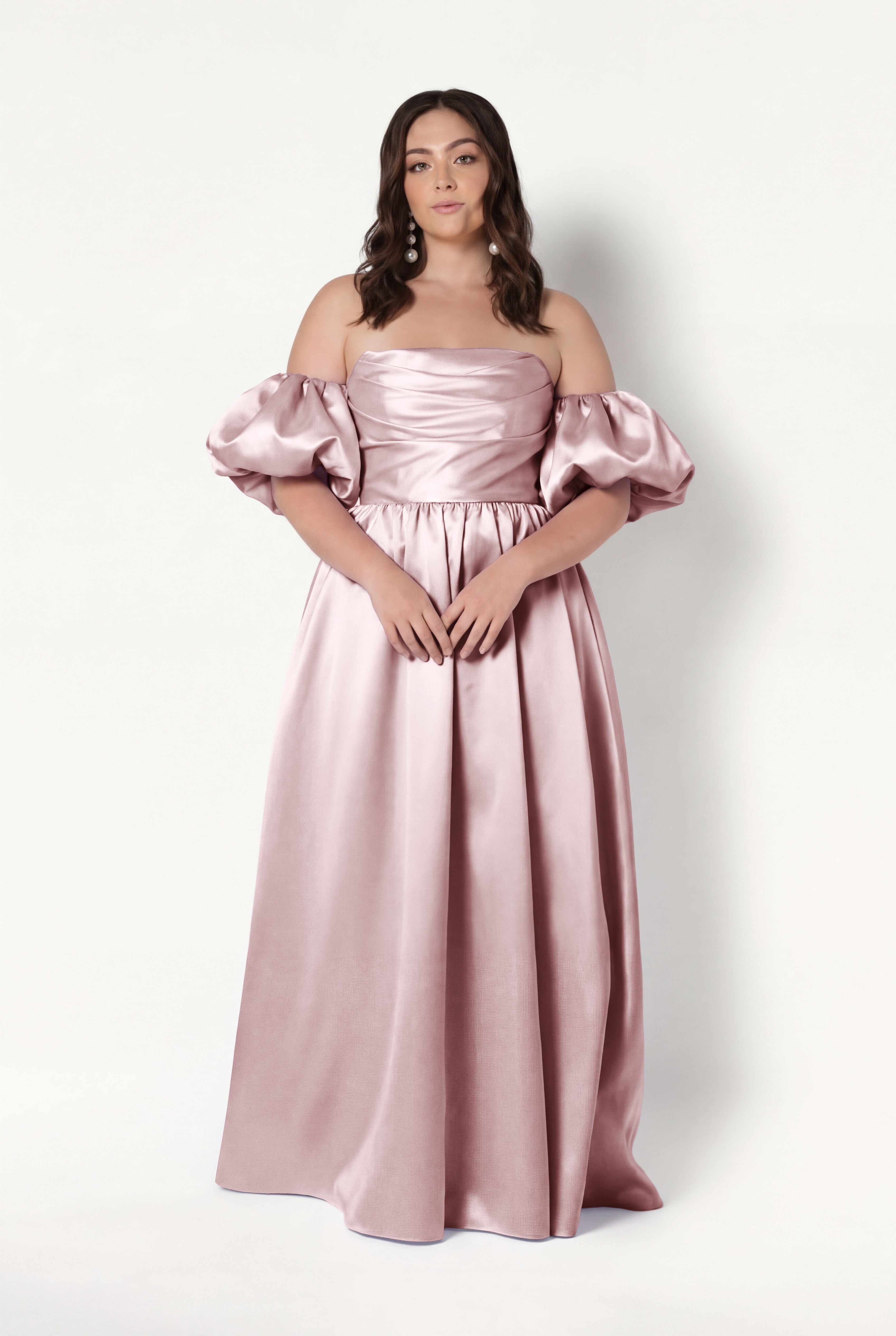 A woman stands against a plain backdrop wearing the Blair Bridesmaid Dress in Smoked Blush Satin Twill by TH&TH—an off-the-shoulder, floor-length gown with detachable puff sleeves. She has long wavy brown hair and a calm expression.