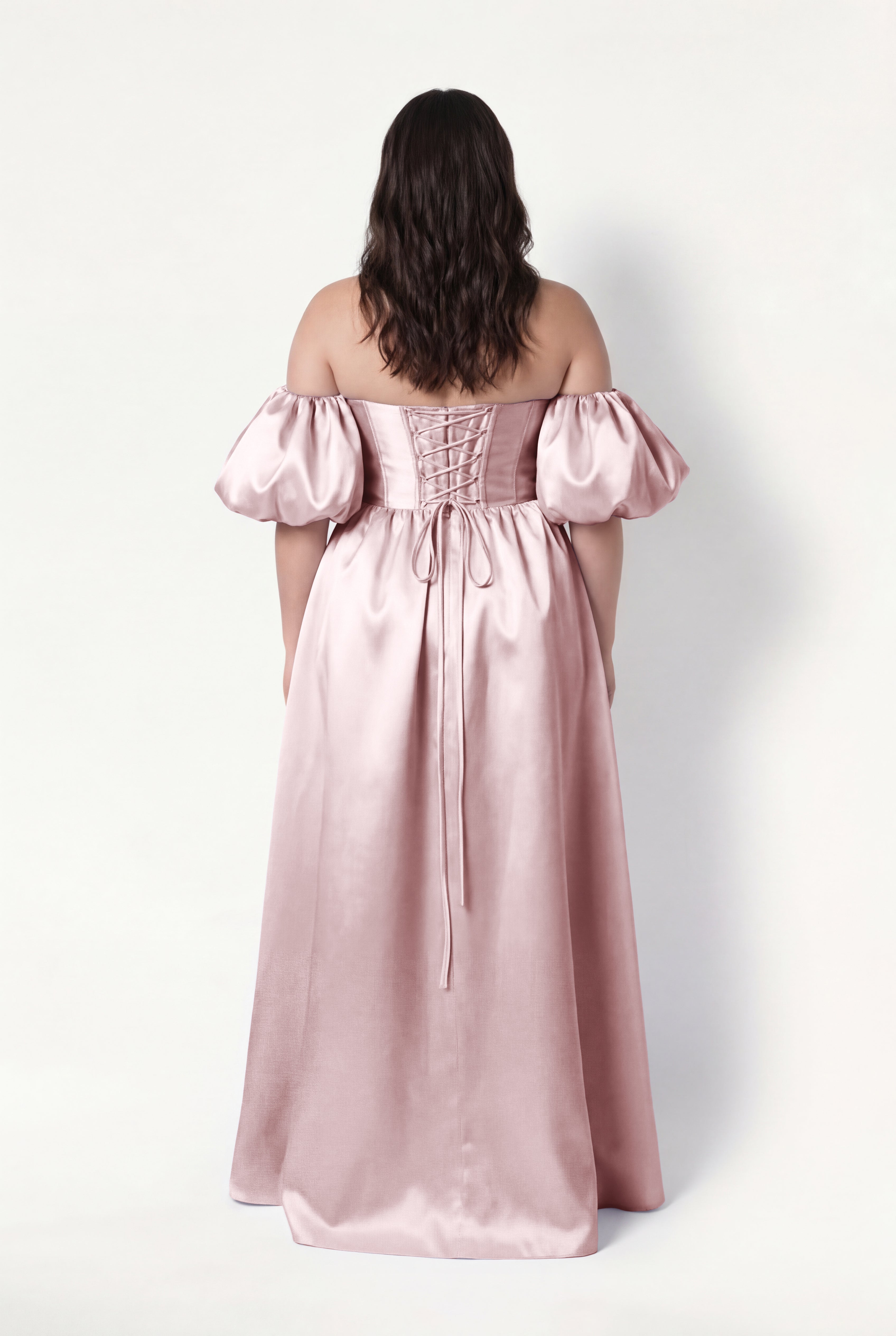 A woman with wavy brown hair faces away, wearing the TH&TH Blair Bridesmaid Dress in Smoked Blush Satin Twill with detachable puff sleeves and a lace-up back, standing against a plain white background.