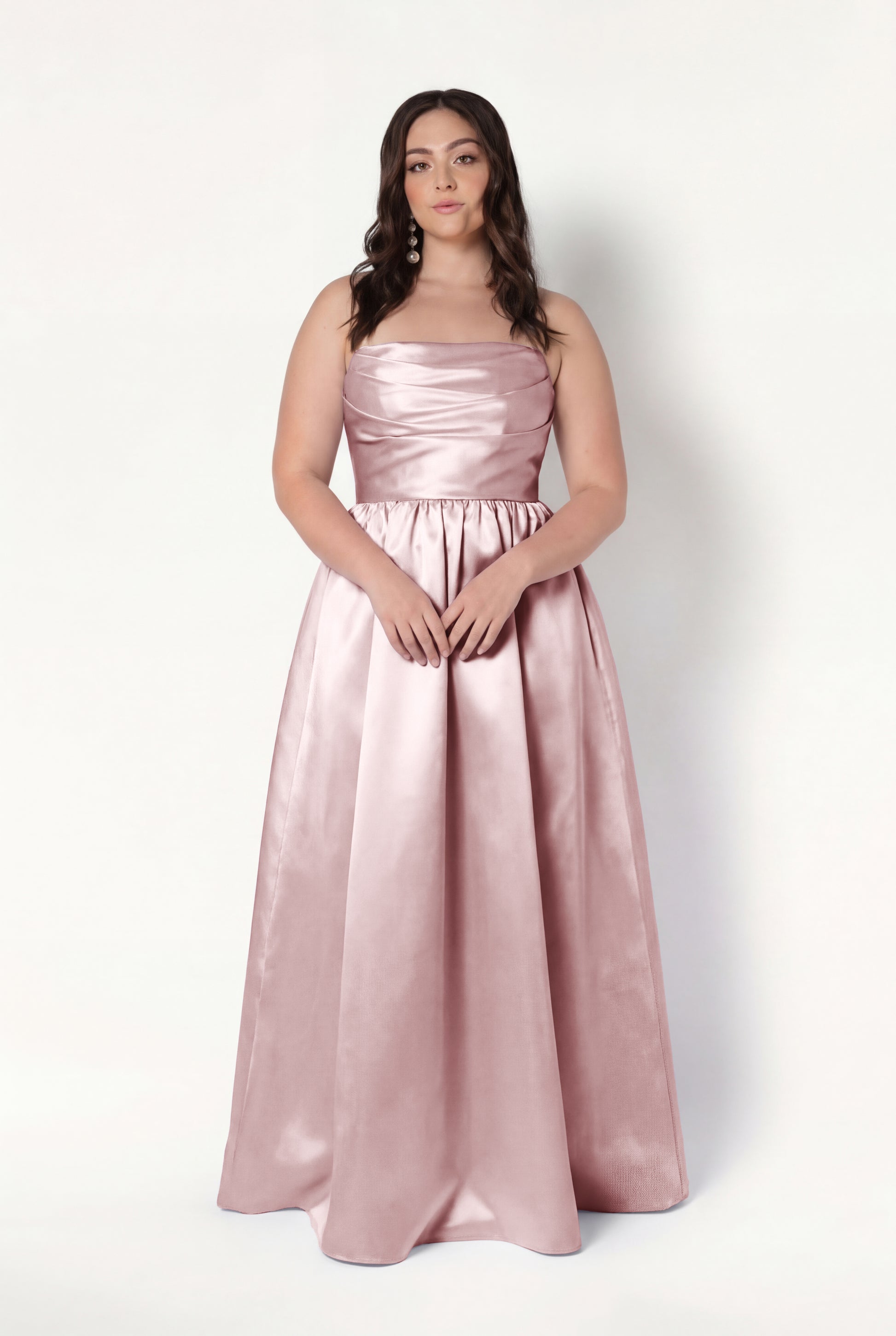 A woman with long, wavy brown hair wears the TH&TH Blair Bridesmaid Dress in Smoked Blush Satin Twill, a sleeveless, floor-length gown with optional detachable puff sleeves. She stands against a plain white background, hands on the skirt.
